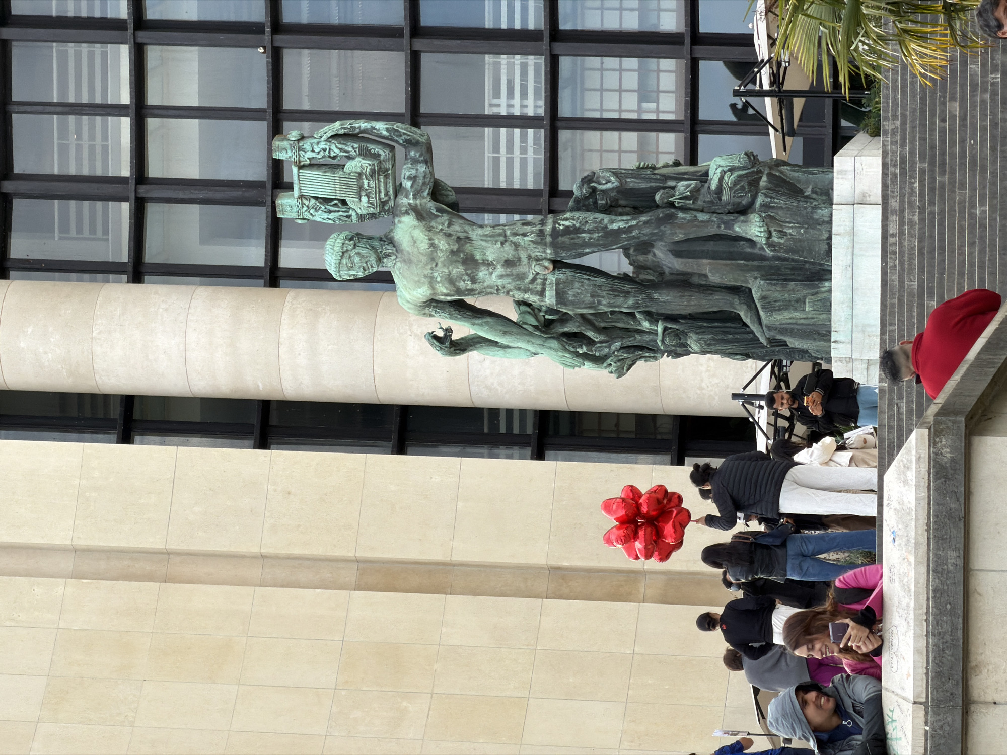 The image depicts a scene in Paris, France, likely at the Trocadéro. A weathered, green-patina statue of a nude male figure, holding a lyre and seemingly emerging from rough terrain, stands in the foreground. Behind the statue, a multi-story building with a light-colored facade and dark-framed windows rises. A group of people is gathered on a staircase leading towards the statue. Some individuals are looking at their phones, while others are standing and talking. A cluster of red heart-shaped balloons is visible amongst the crowd. A portion of palm fronds is seen above the staircase.