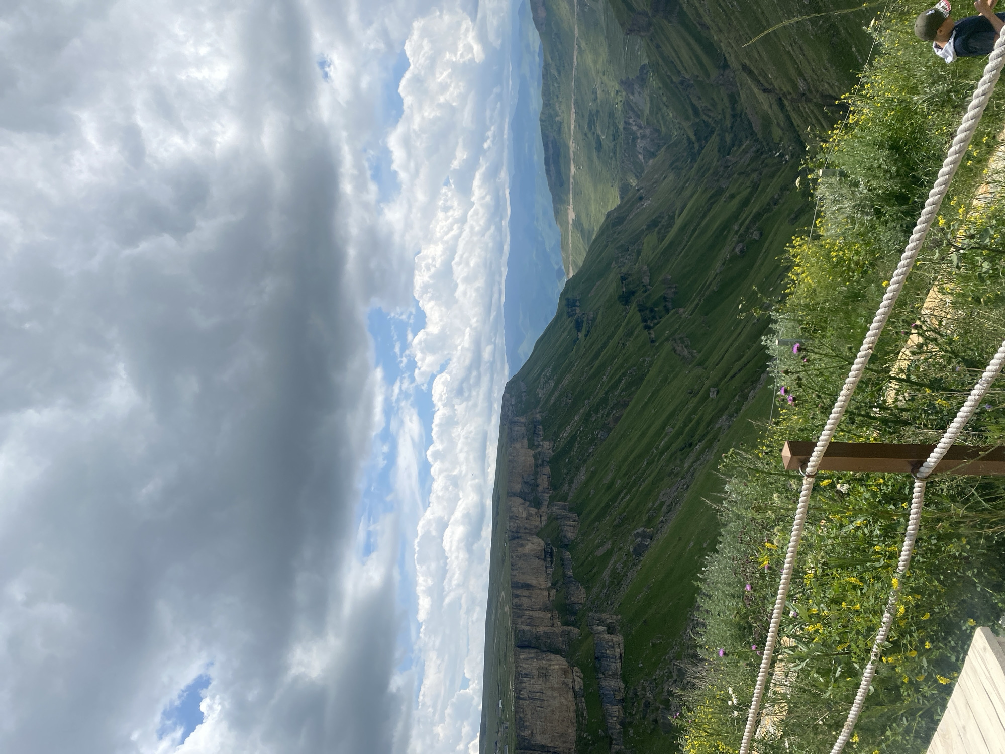 This is an outdoor, full-frame, eye-level shot of a vast mountain landscape under a cloudy sky. The foreground features a wooden boardwalk and a rope railing bordering a steep, green mountainside covered in wildflowers, including yellow and purple varieties. In the bottom right corner, a young person wearing a dark t-shirt and a baseball cap is visible. The middle ground showcases the dramatic, steep slopes of a canyon, carved into the green terrain. The background reveals layered mountain ranges fading into the distance. The scene is likely the famous Khunzakh plateau or a nearby canyon in Khunzakh, Russia, known for its breathtaking natural beauty.
