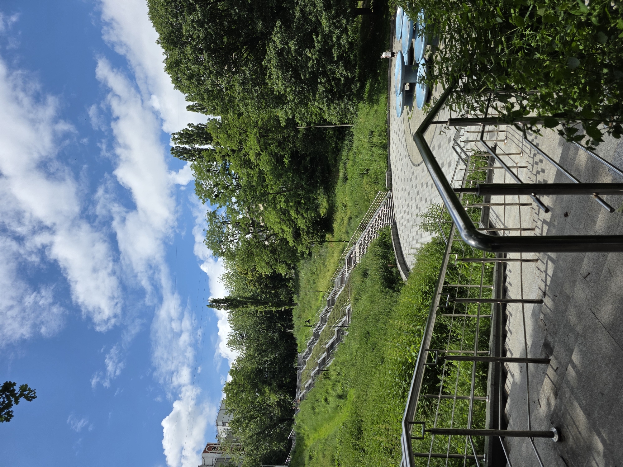 The image captures a bright, sunny day in Kyiv, Ukraine. The scene is an outdoor staircase winding down a lush, green hillside covered in dense vegetation. The sky is a vibrant blue, dotted with fluffy white clouds, suggesting fair weather. Tall, leafy trees line the top of the hillside, providing ample shade.
The staircase is made of stone with metal railings, appearing to be part of a park or a public pathway. The overall impression is one of a well-maintained public space designed for recreation or a scenic route. In the foreground, the metal railings of the staircase are prominent, reflecting the sunlight. Further down the slope, multiple flights of stairs are visible, separated by grassy areas. A person in a white shirt and dark shorts is visible near the top of the staircase, standing on a paved area with blue circular objects, possibly part of a decorative installation or playground equipment.
The scene evokes a feeling of tranquility and natural beauty, enhanced by the clear blue sky and abundant greenery. The presence of the staircase and the paved area suggests a developed urban park. The time of day appears to be midday or early afternoon due to the bright lighting and the shadows cast. No visible text is present in the image.