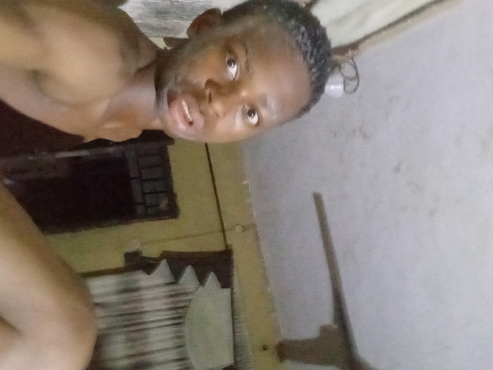 The image shows a person with dark skin, short hair, and light-colored eyes looking upwards at the camera. The person is bare-chested, and the background includes a window with bars, a light-colored wall, and some other indistinct objects. The location is Nkpor, Nigeria.