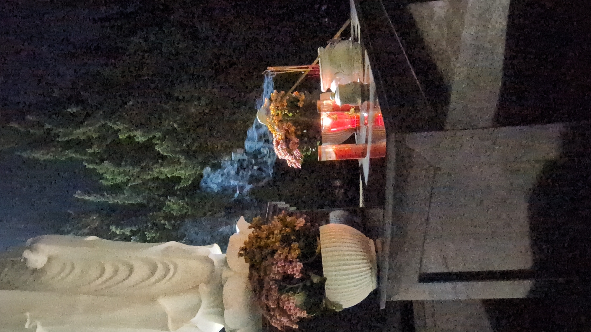 A nighttime shot of a Buddhist shrine in Da Lat, Vietnam, rotated ninety degrees clockwise. The bottom left quadrant shows a section of a white, ornate stone statue, possibly depicting Buddha. To the right of the statue, a white urn or pot sits atop a stone base, overflowing with pink and orange flowers. Above the flowers, rising in wisps, is the white-blue smoke from burning incense. Further up, a red lantern in a white holder hangs suspended by metal supports. The background shows foliage in the darkness. The rightmost quadrant of the image shows a grey cement or stone wall. The overall impression is a serene religious site at night.