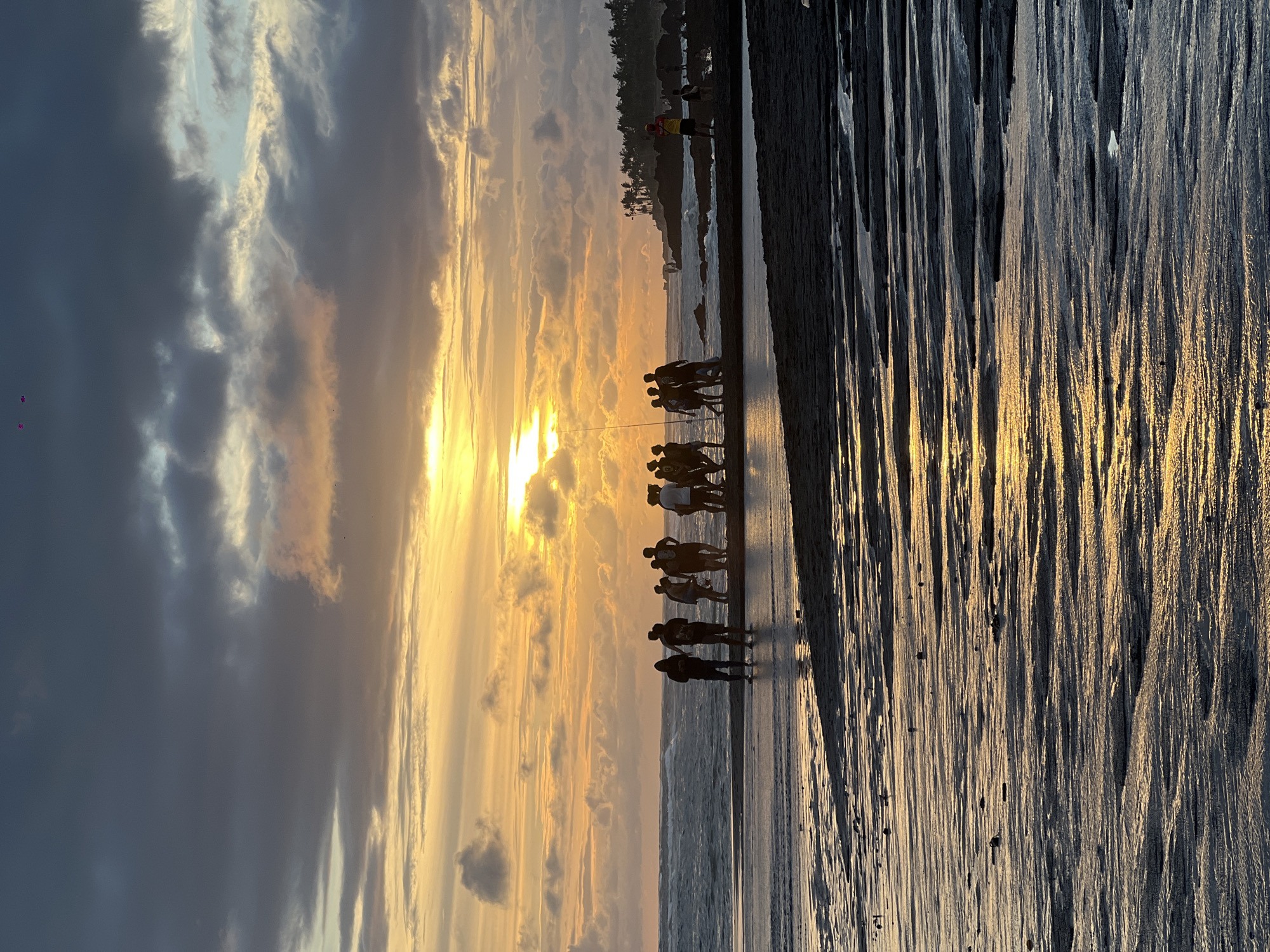 The image captures a coastal scene in Mengwi, Indonesia, at sunset. A group of about ten people, silhouetted against the setting sun, are standing or walking on the wet sand near the water's edge. Their shadows stretch long across the wet sand. Behind them, the ocean meets the shore, and further back is a darker landmass with trees and some indistinct figures, possibly including a lifeguard. The sky is filled with clouds, with the sun creating a bright glow and casting a golden hue across the scene. The wet sand reflects the sunset colors, enhancing the scene's visual impact. The texture of the sand is visible, with patterns created by the receding tide.