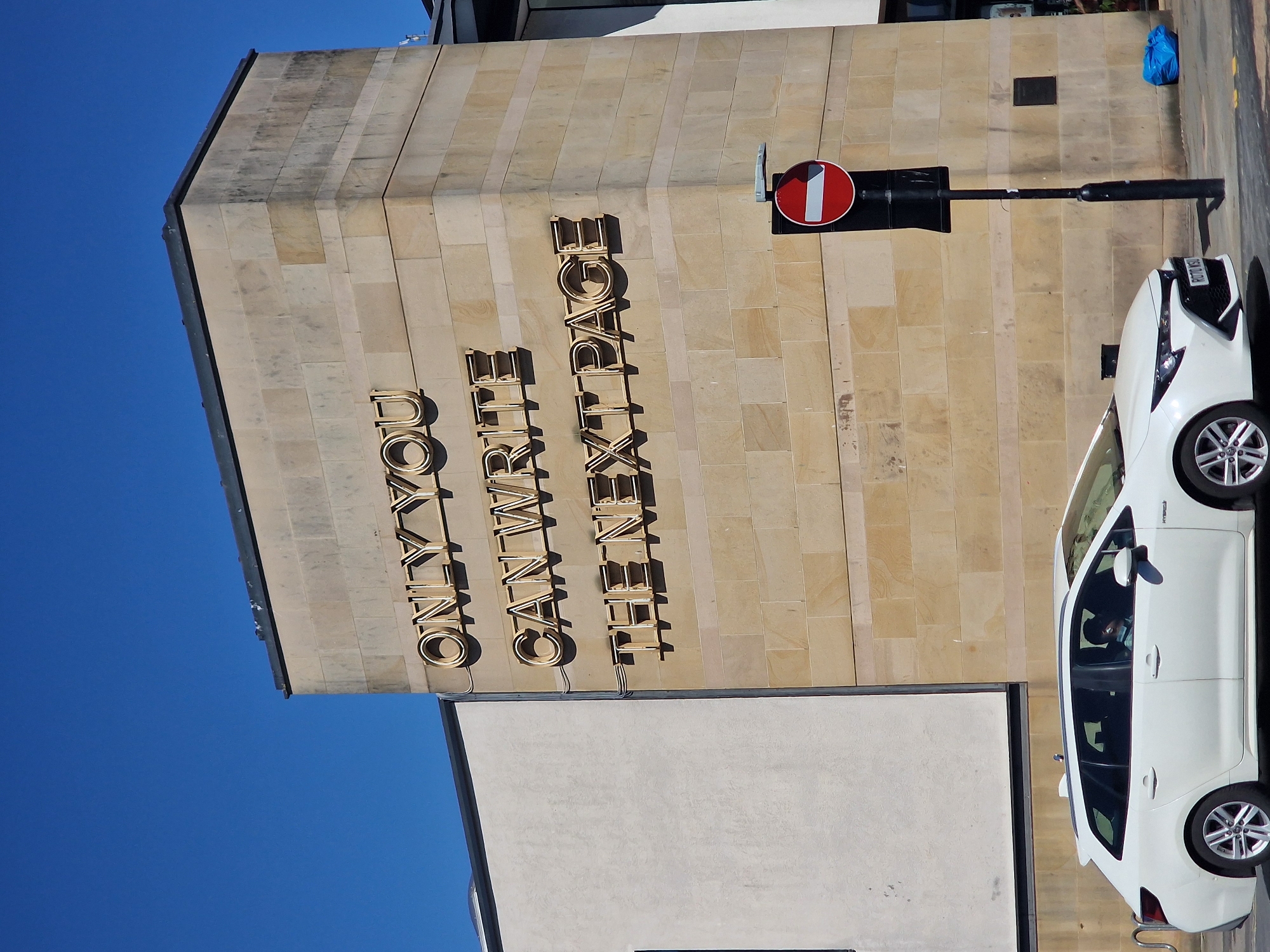 Here is a description of the image:
The image shows the exterior of a building in Durham, United Kingdom, identified by the gold lettering as "ONLY YOU CAN WRITE THE ENTRANCE." A white car is parked adjacent to the building on the right-hand side of the frame. A black and red "no entry" sign is mounted on a pole fixed to the building, beside the car. The building is constructed from light-colored stone bricks. The sky is a clear blue.