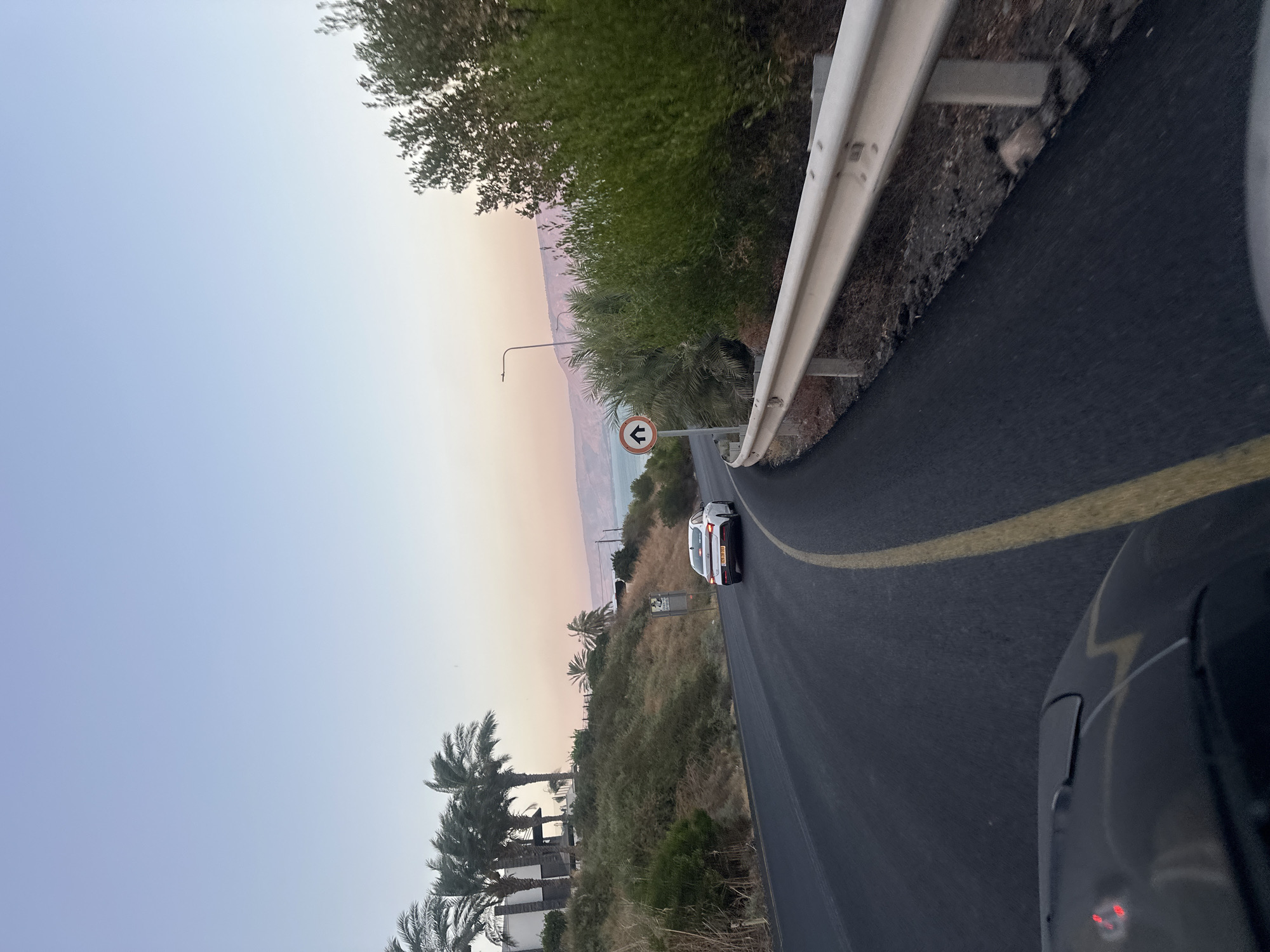 The image depicts a road winding through a landscape with a body of water in the distance. The road is paved with a yellow line marking the lane. On the left side of the road, there is a hillside covered with vegetation and several palm trees. A white car is driving away from the camera on the road. In the background, there is a body of water, possibly the Sea of Galilee, and hills. The sky is clear and shows a gradient of colors from light blue to pink, suggesting it is either sunrise or sunset. A sign with a circular orange border and a black house icon is visible on the side of the road. On the right side of the road, there is a white metal guardrail and dense green foliage. The foreground shows a portion of a dark-colored vehicle's hood and windshield wipers, indicating the photo was taken from inside a car. The location is identified as the Emek HaYarden Regional Council in Israel.