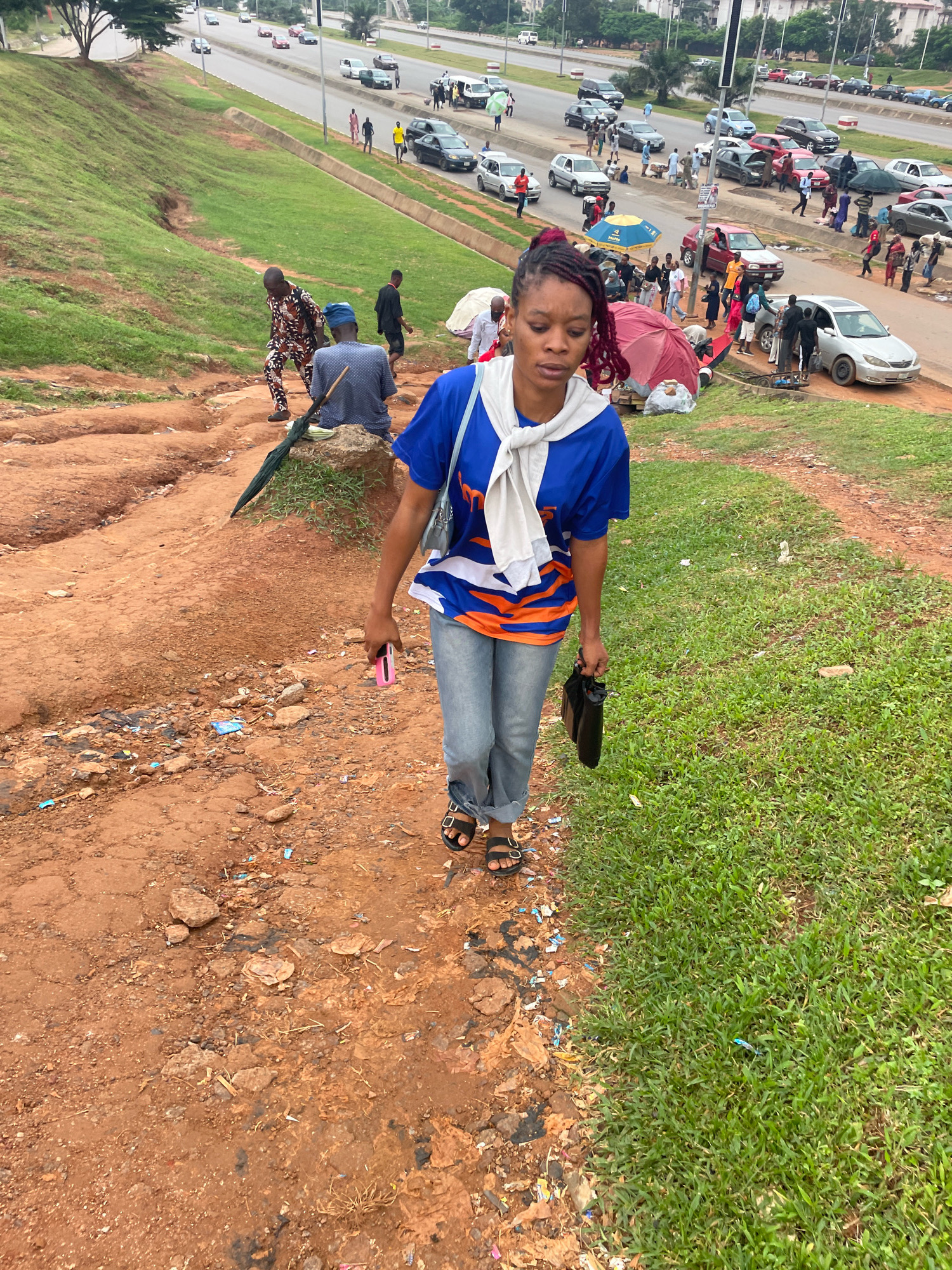 In Abuja, Nigeria, a woman with braided hair dyed red walks down a steep, eroded dirt path adjacent to a grassy embankment. She wears a blue t-shirt with an orange and white design and a white scarf tied around her neck. Her attire also includes denim jeans and black sandals. In her right hand, she holds a pink cell phone, and in her left, a black bag. Behind her, other individuals are visible on the path and on the grassy area. The background reveals a busy road with numerous cars and people, suggesting an urban setting. The environment is characterized by uneven terrain, sparse vegetation, and scattered debris on the path.