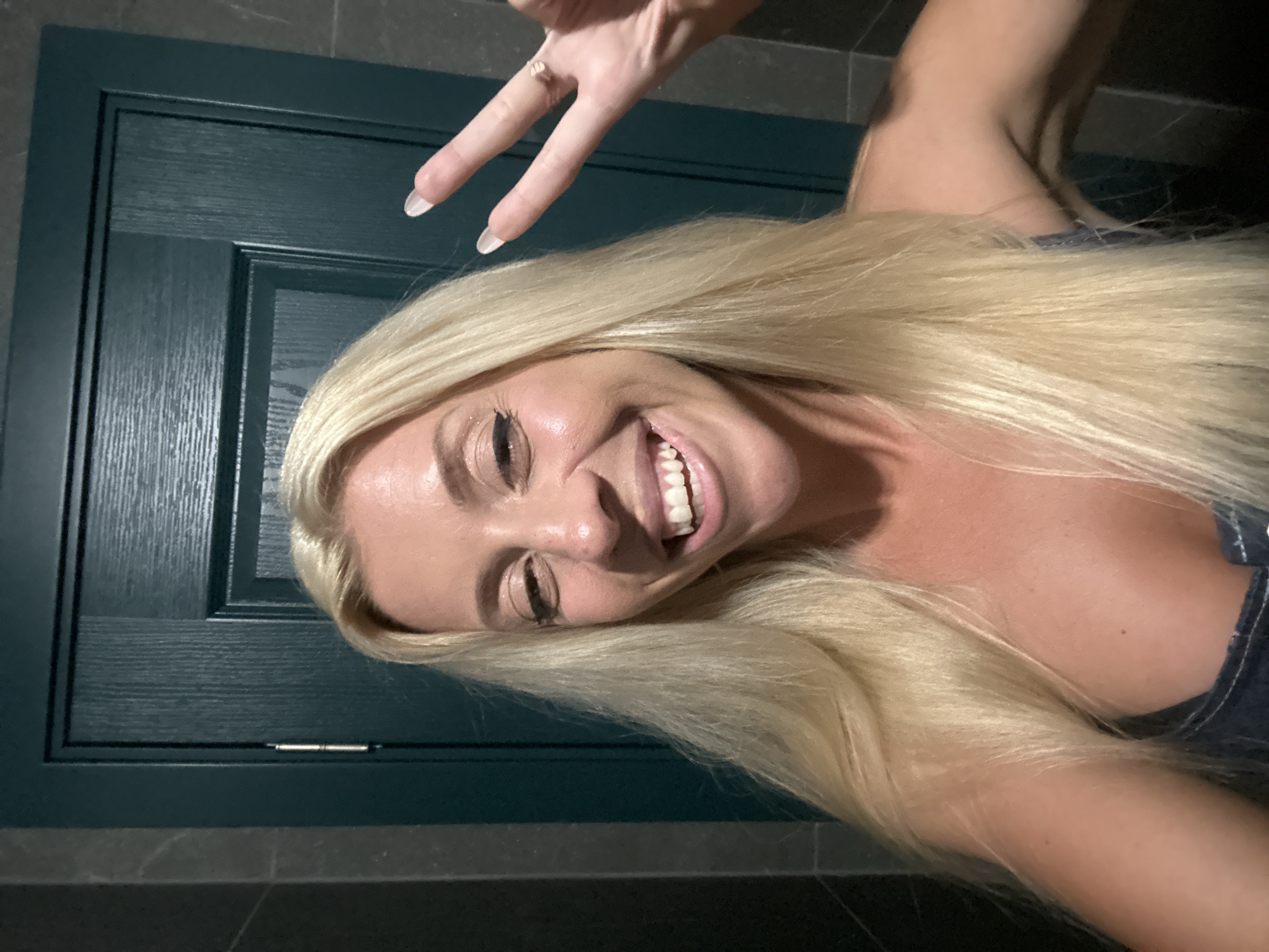 A woman with blonde hair and a bright smile is taking a selfie. She is wearing a dark blue top. She is making a peace sign with her right hand, and a ring is visible on her ring finger. She has on winged eyeliner and appears to be wearing makeup. Behind her is a dark green door with a wood grain texture. The walls on either side of the door appear to be grey tile. There are no identifiable landmarks in the image that would indicate the location of Pazardzhik, Bulgaria.