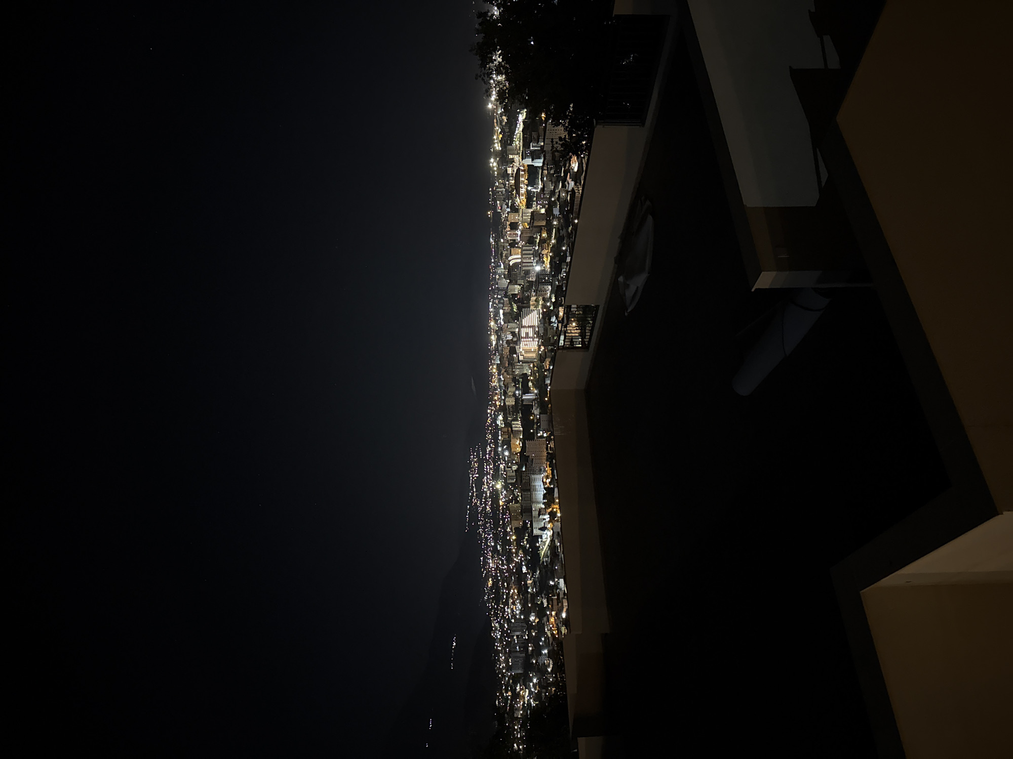 The image shows a night-time view of a city from an elevated vantage point, possibly a balcony. The cityscape is illuminated by numerous lights, suggesting a dense urban area. The city stretches along a coastline and inland. A dark area to the left might represent the sea. In the foreground, architectural elements of a building, such as walls and eaves, are visible. The location is likely Šušanj, Montenegro, as it matches the coastal, hilly terrain with concentrated urban development. No people are visible.