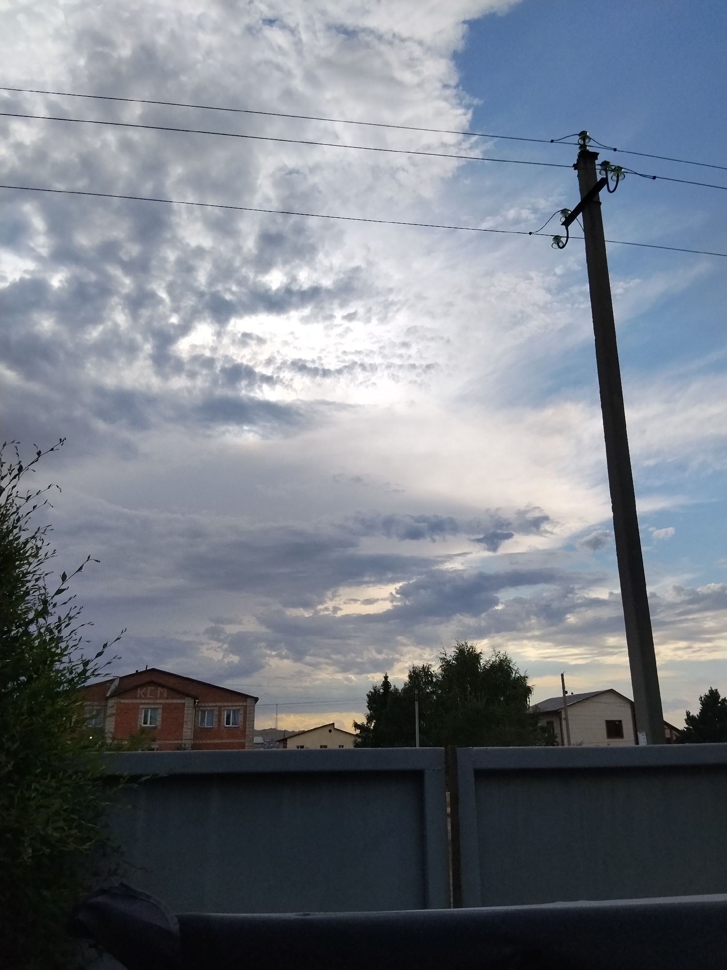 A view of a cloudy sky with a prominent utility pole on the right side. The pole is made of concrete and supports overhead power lines with ceramic insulators. In the middle ground, there are several buildings visible, including a brick house with windows and the letters "KEN" on its facade, and other residential structures. A dark metal fence runs across the foreground. To the left, the branches of a green leafy bush are visible. The overall scene suggests an outdoor residential area.