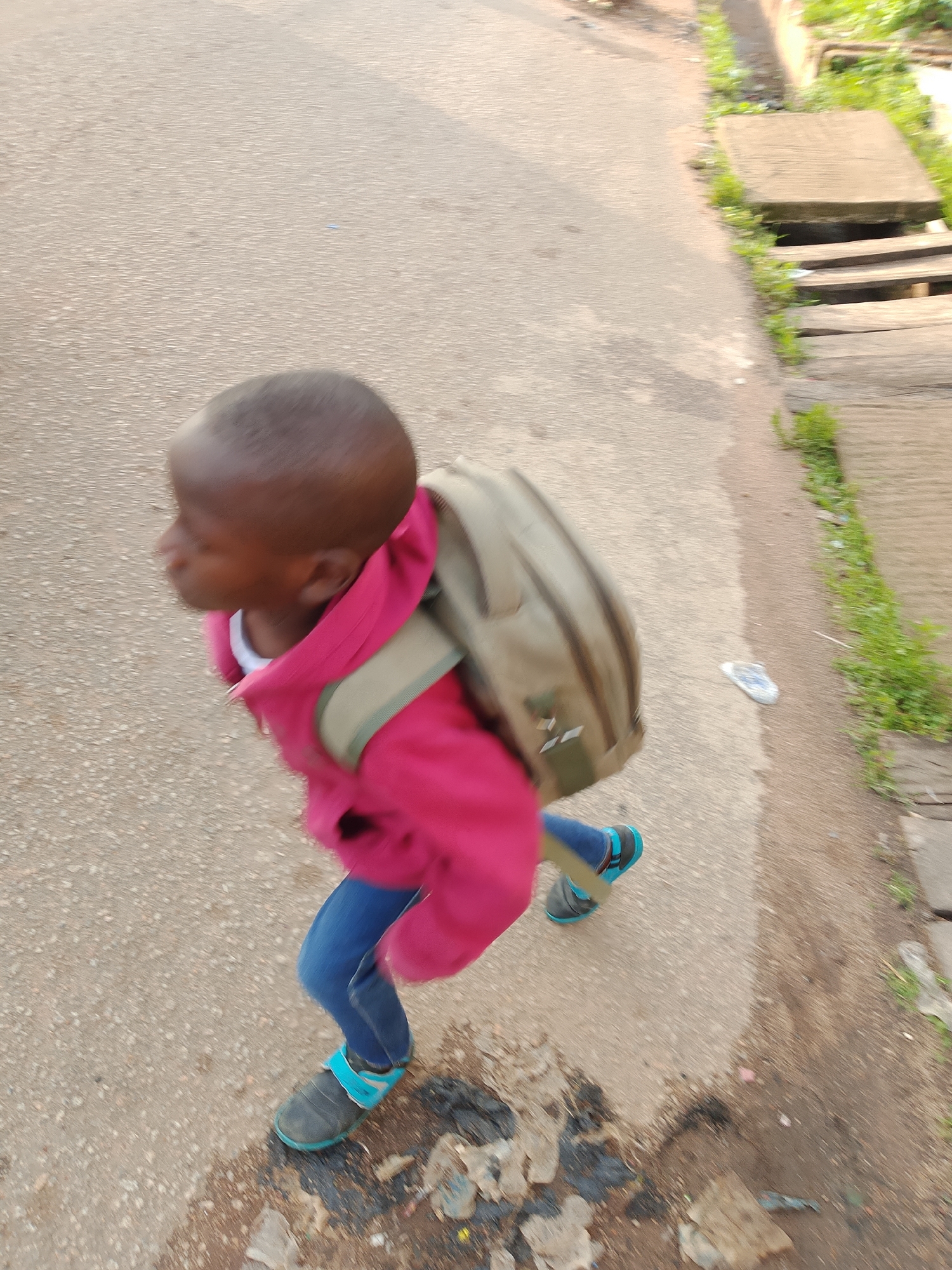 In Jos, Nigeria, a young child with a shaved head is captured in motion, walking on a paved surface. The child is wearing a bright pink hooded jacket and blue jeans, with blue and black shoes. A light brown backpack is slung over the child's shoulders. The background features a drainage ditch with green vegetation growing along its edge, and a wooden structure that appears to be a bridge or walkway. The ground is a mix of concrete and dirt, with some scattered debris visible. The image is taken from a high angle, giving a dynamic perspective of the child's movement.