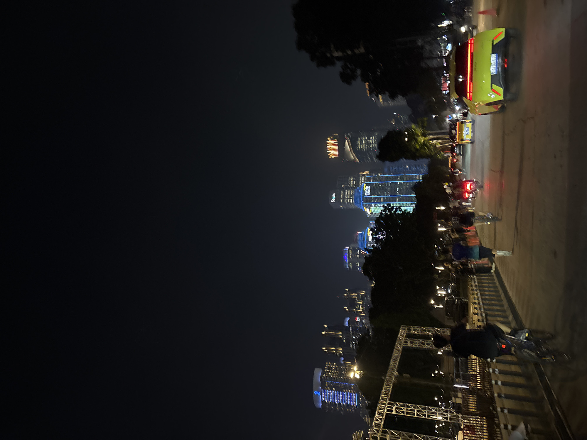 A vibrant urban night scene captures a bustling street and skyline in Central Jakarta, Indonesia. The image, presented as if rotated 90 degrees clockwise, displays a dynamic cityscape illuminated by numerous artificial lights against a dark sky.
In the foreground, a large metal truss structure stands prominently on the left side, potentially for an event or signage. Along the right, a multi-lane road is active with traffic. A bright, light green or yellow car with distinct red taillights is visible, displaying an Indonesian license plate starting with "B 17xx Uxx" (e.g., B 1740 UQQ). Behind it, other vehicles, including what appears to be another car or taxi and several motorbikes with illuminated taillights, are part of the night-time flow.
Adjacent to the road, a barrier separates the traffic from pedestrians and cyclists. Several individuals are present, with one person in dark clothing and a helmet clearly riding a bicycle in the lower right portion of the image. Other figures are also visible further along the street, some on bicycles and others walking.
The background is dominated by a dense array of modern skyscrapers and tall buildings, their numerous windows and exterior lighting brightly illuminating the scene. Many structures feature unique architectural lighting, with some displaying prominent blue light patterns. Text is discernible on several buildings: "Sarinah" and "Sequis" are clearly legible on different structures, identifying specific locations. Dark trees with dense foliage are scattered throughout the scene, partially obscuring some of the illuminated buildings. The overall impression is one of a lively, modern metropolitan area at night.
