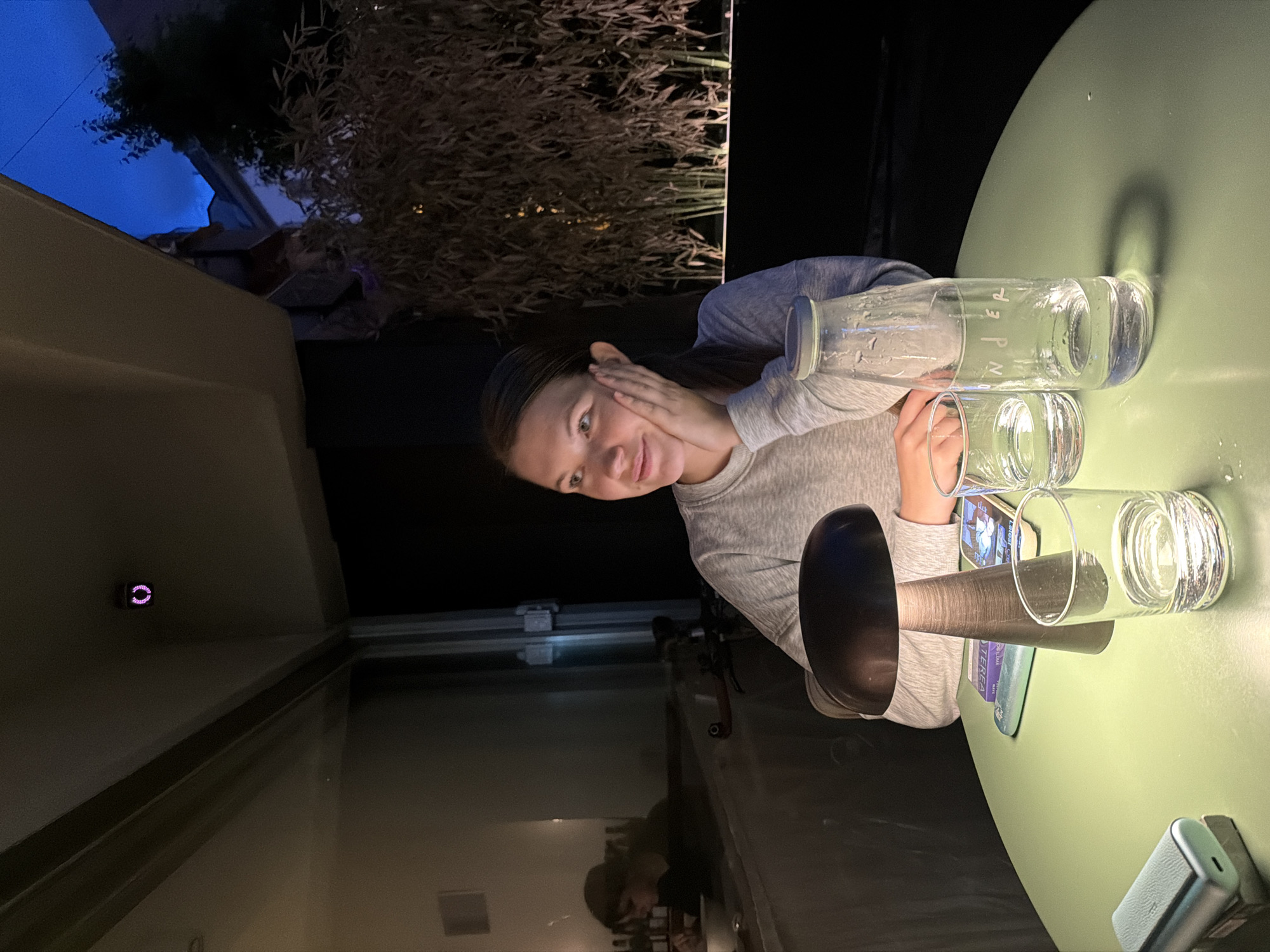 The image presents a scene at what appears to be an outdoor dining area in Belgrade, Serbia. A woman is the focal point, seated at a table. She is wearing a light gray long-sleeved shirt and has her hand near her face, giving the impression of posing for the camera.
On the table, there are several water glasses, a glass bottle with a gray lid, a black candle holder, and at least two books. A mobile phone and possibly a charging case are also on the table. The table has a light green surface.
In the background, there are buildings and trees. There is also a window, possibly reflecting the light from the sky. The lighting suggests it is either evening or nighttime. There is also an indoor section that has reflected objects on the glass windows. The city location is identified as Belgrade, Serbia.