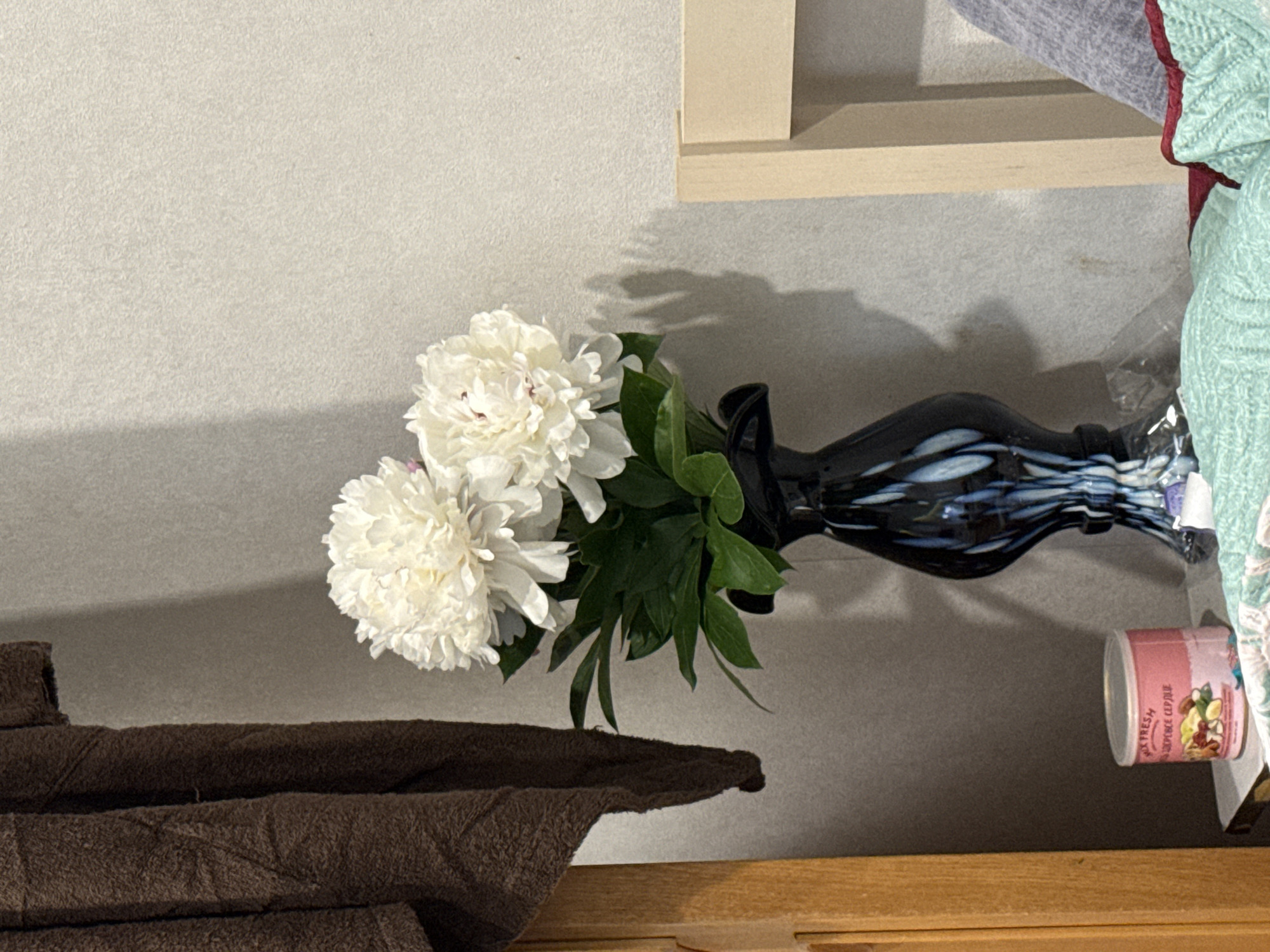 The image depicts an indoor still life with a vase of white peonies on a bedside table. To the left, a brown, fuzzy towel or garment is draped over a wooden surface, possibly a headboard. In the center, a tall, dark vase with blue and white swirls holds a bouquet of lush white peonies with green leaves. To the right, a portion of a light-colored wooden chair is visible. On the table below the vase, a small, pink tin with a lid contains what appears to be a mix of nuts and dried fruit, with some text in Russian. The background is a textured, light-colored wall. The overall scene suggests a domestic setting, likely a bedroom, in Valday, Russia.
