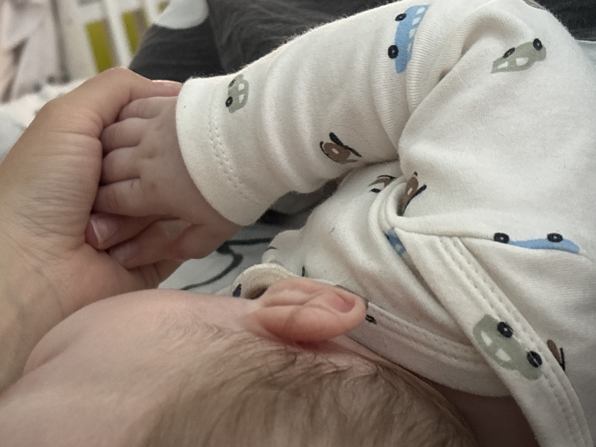 The image shows a close-up view of an adult holding a baby. The baby is lying down, and the adult is holding the baby's hand. The baby is wearing a white onesie with car patterns. The focus is on the baby's ear and hand, along with the adult's hand holding it. No information suggests it is taken in Раздолье, Russia.