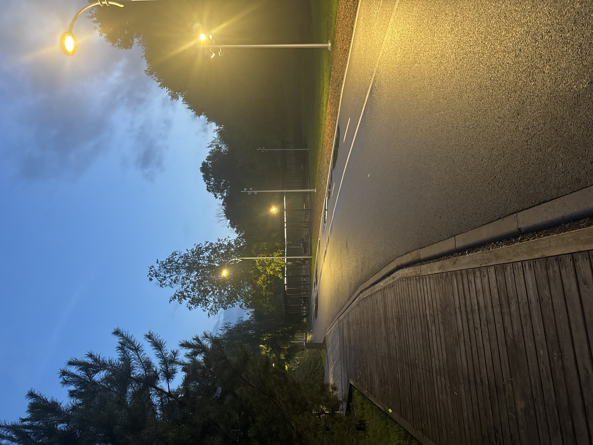 The image shows a paved road next to a wooden walkway in Ufa, Russia, illuminated by streetlights during twilight. The sky is a deep blue with some wispy clouds. Trees line the sides of the road and walkway, with a covered seating area visible in the background. The road surface appears wet, reflecting the yellow glow of the streetlights. The wooden walkway is made of horizontal planks and is slightly elevated from the surrounding grass. There are no people or animals visible in the scene.