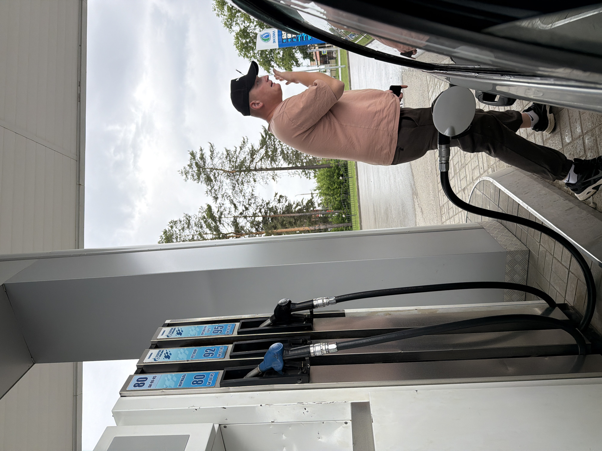 The image depicts a man refueling a dark-colored car at a gas station in Chaikovsky, Russia. The man stands beside the vehicle, with the fuel nozzle inserted into the car's tank. He wears a black baseball cap, a light peach-colored short-sleeved t-shirt, dark pants, and black and white sneakers. He is looking upwards and appears to be speaking or gesturing with one hand near his face.
The scene is set outdoors on an overcast day, with trees visible in the background against a cloudy sky. A white gas pump features multiple nozzles and displays different fuel grades with Cyrillic text. Visible labels include "80 НОРМАЛЬ" (Normal 80), "80 AW-80-K5", "92 РЕГУЛЯР" (Regular 92), and "95 ПРЕМИУМ ЕВРО" (Premium Euro 95). One of the nozzles, specifically for 92 octane, is inserted into the car. In the background, a sign for "Экоил" (Ecoil), likely the gas station brand, is visible, along with price displays such as "92 63.9" and "95 69." The ground is paved with light-colored bricks or concrete.
