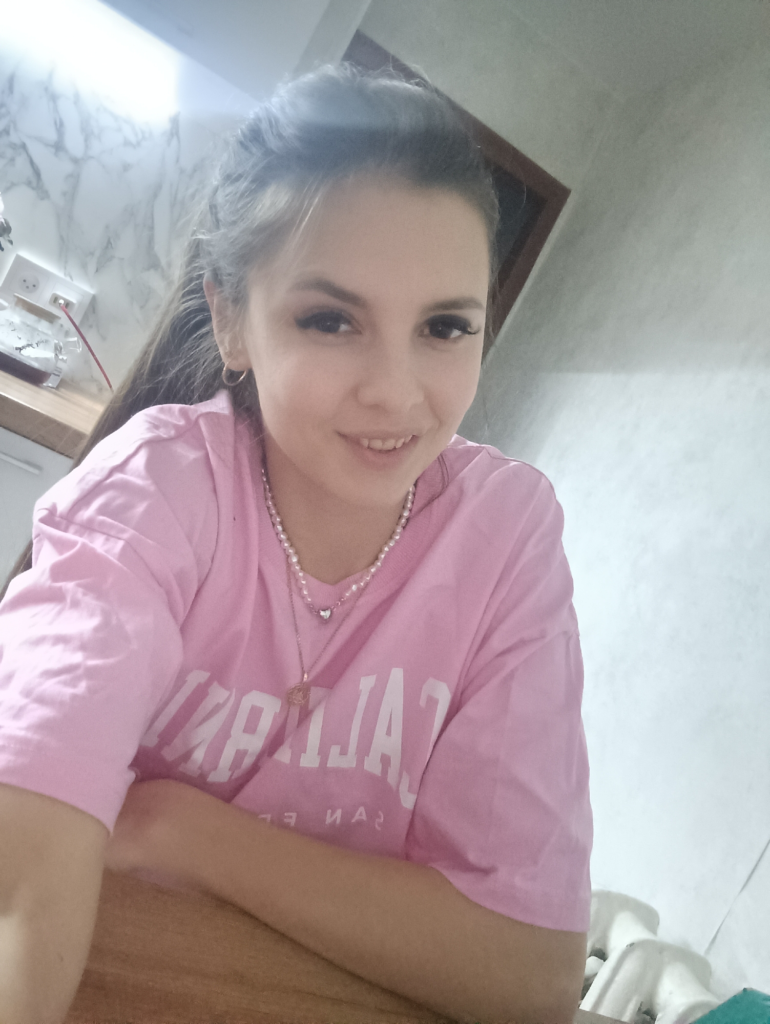 The image shows a young woman wearing a pink t-shirt with white lettering, a pearl necklace, and a gold necklace with a heart pendant. Her hair is pulled back, and she is smiling. She appears to be sitting at a wooden table. In the background, there is a marble-patterned wall, an electrical outlet, and a framed picture. The location is in the city of Unknown, Unknown.
