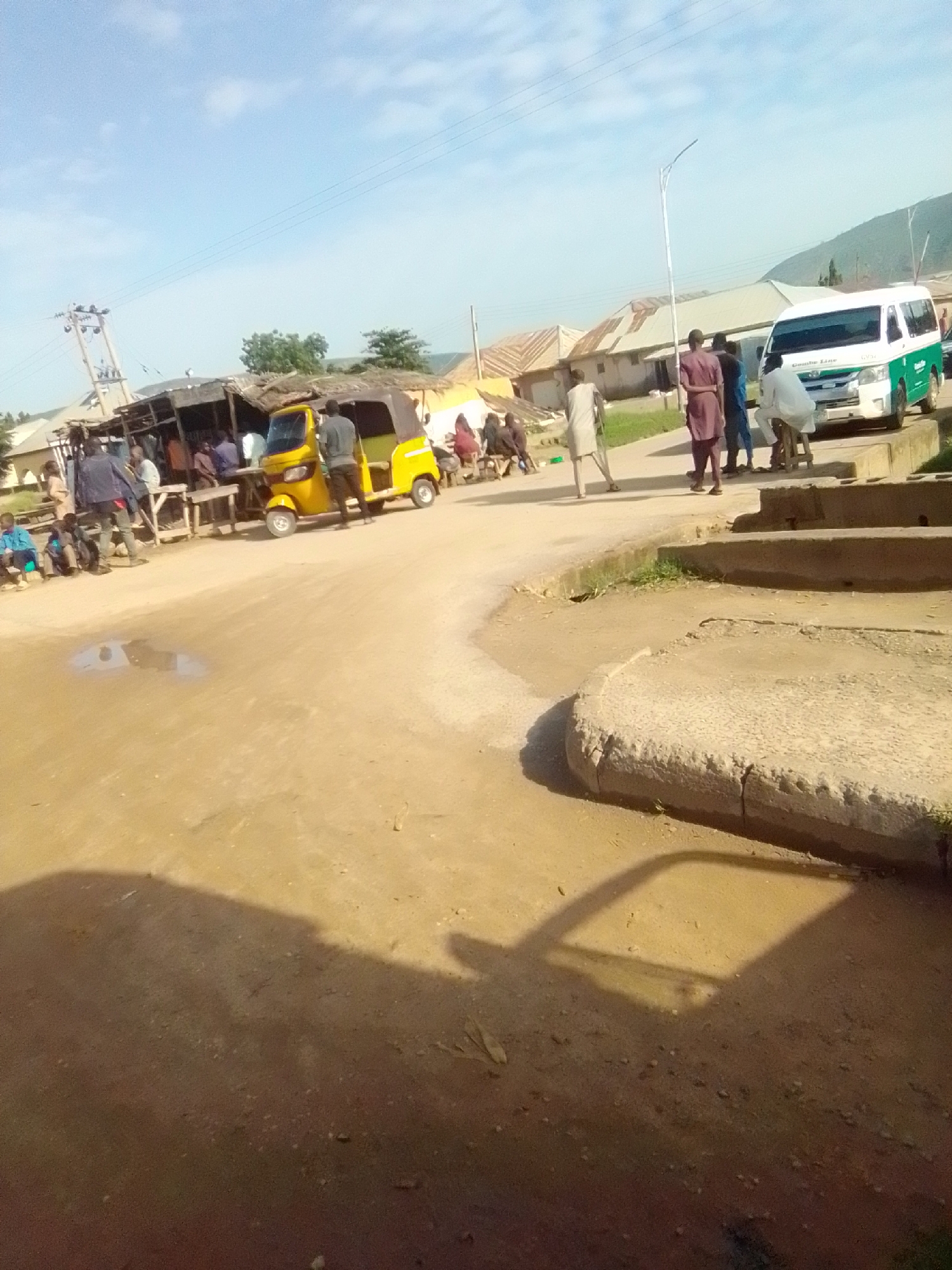 In Kwadon, Nigeria, a wide, unpaved street is the scene. In the middle of the street, a yellow auto-rickshaw is parked. To the right of the auto-rickshaw, a white van with green accents is parked. Several people are standing and sitting around the auto-rickshaw and van. Buildings with corrugated metal roofs are visible in the background. The sky is mostly clear with some clouds. The ground is dirt and dusty, with a few puddles of water. The overall impression is of a busy, informal market or transportation hub.