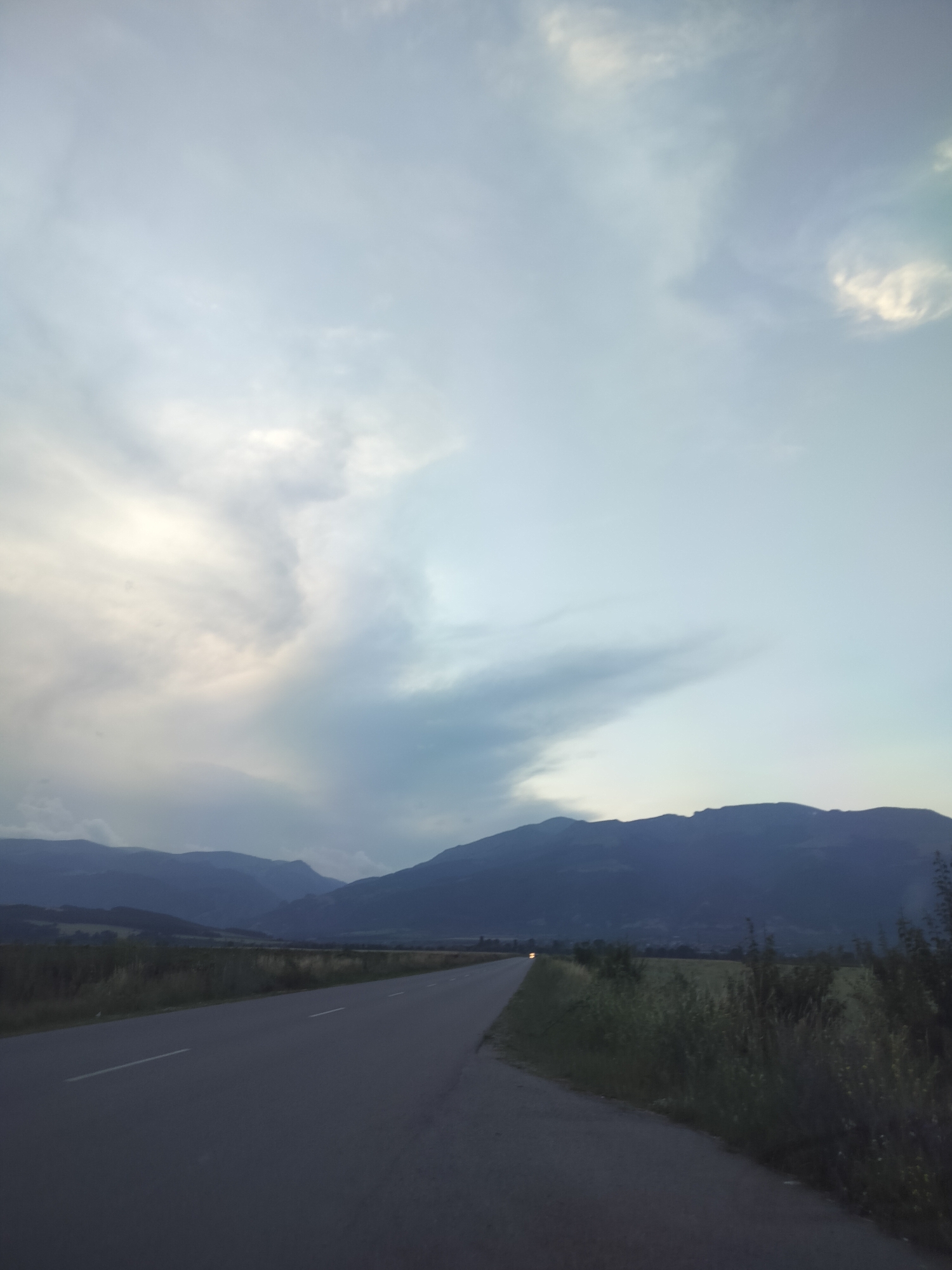 The image depicts a rural scene in Manolovo, Bulgaria, at dusk. A paved road stretches into the distance, flanked by grassy fields and low-lying vegetation. In the background, a range of dark, silhouetted mountains forms the horizon. The sky is vast and cloudy, with dramatic formations of white and grey clouds that create a sense of depth and atmosphere. The overall mood is serene and somewhat dramatic, possibly due to the approaching or receding storm suggested by the cloud formations. The lighting indicates that it is either early morning or late evening, with the sun likely low on the horizon. There are no people or specific objects visible besides the road and natural landscape. No text is present in the image.