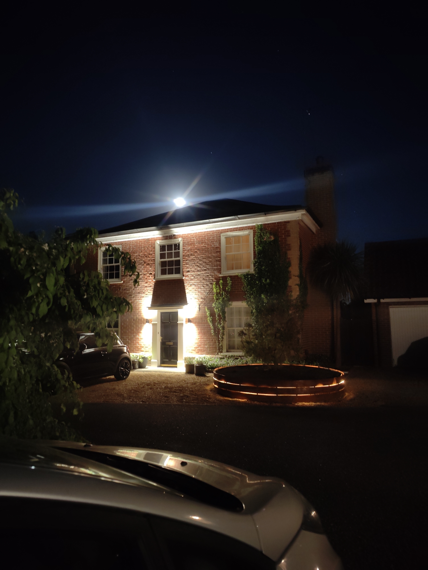The image captures a brick house at night, brightly illuminated by artificial lights under a dark sky. The two-story house features multiple white-framed windows and a dark front door, flanked by two wall-mounted sconce lights. A prominent, bright light source, possibly a floodlight, is positioned above the front of the house, creating a significant lens flare across the upper portion of the image.
In the foreground, the hood and windshield of a light-colored car (likely silver or white) are visible on the bottom edge, partially obscuring the driveway. Further up the driveway, to the left of the house, a dark-colored Mini Cooper is parked, partially hidden by dense green foliage. To the right of the driveway, a large, circular planter or raised bed with an internal orange glow adds warmth to the scene. The house itself has ivy growing on its walls and a slender tree to the right of the front door. A chimney is visible on the right side of the house, and beyond it, a dark building, possibly a garage, with a white door can be seen. The sky is a deep blue-black, with a few faint light specks suggesting distant stars or atmospheric phenomena.
The scene depicts a quiet nighttime setting, with no people visible or specific activity taking place. The artificial lighting strongly contrasts with the dark surroundings, highlighting the architectural features and landscaping of the property. There is no visible text in the image.