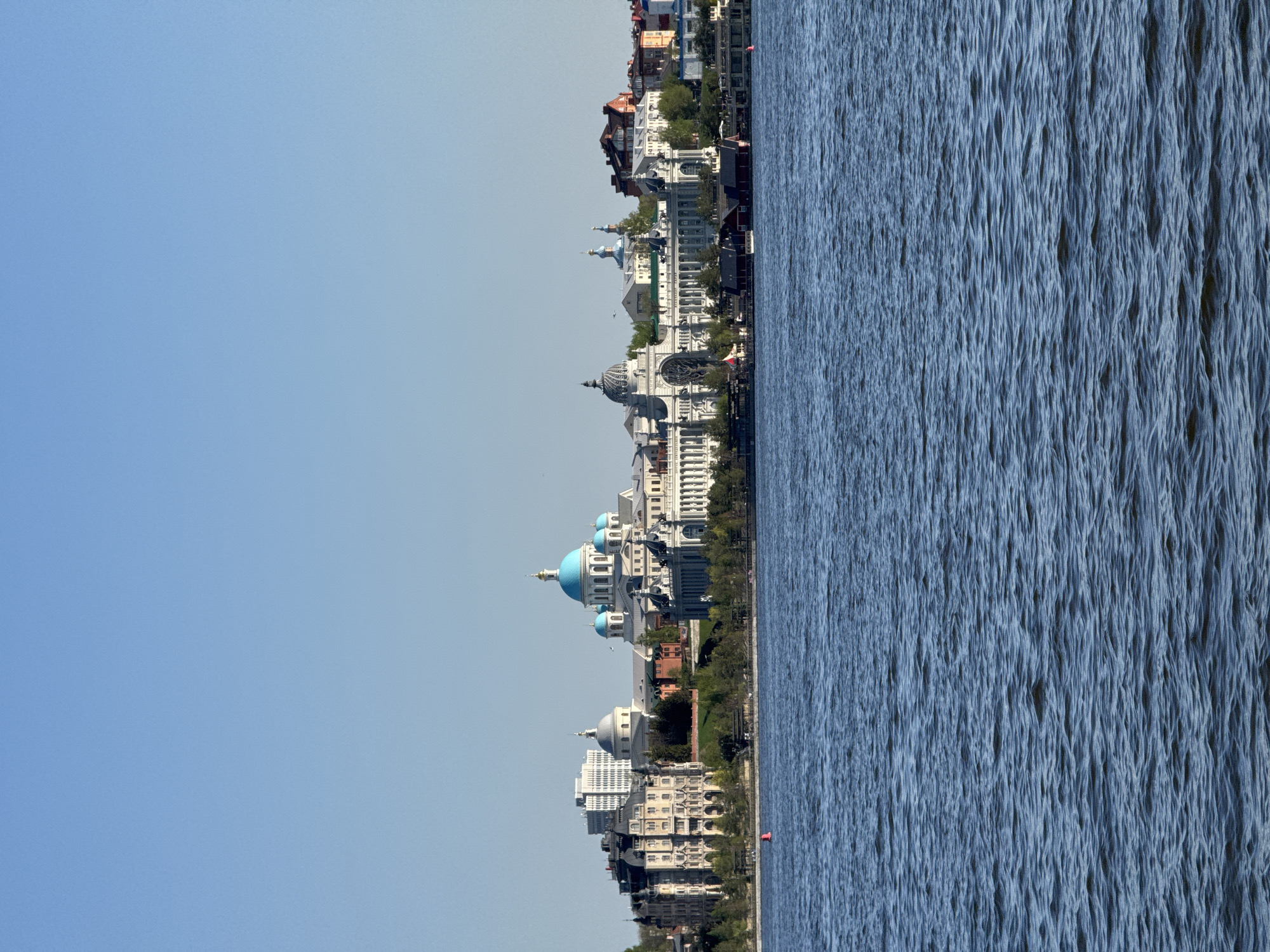 The image depicts the cityscape of Kazan, Russia, oriented vertically with the blue sky dominating the left half and a body of water filling the right half. A series of buildings line the shore, a mix of white structures and domes, plus older buildings with red or brown roofs, and a modern white highrise. Some of the buildings have prominent blue domes. Trees line the space between the buildings and the water. The water's surface is textured with ripples. A single red marker buoy is visible near the shore at the bottom of the frame.