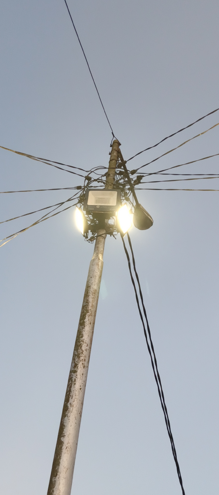 A low-angle shot captures a utility pole with multiple electrical wires and two lights against a clear, pale blue sky. One light, rectangular and modern, is positioned centrally on the pole and is brightly illuminated, casting a strong glow. To its right, a smaller, curved light fixture is also visible. The pole itself appears weathered and worn, with patches of discoloration and texture. The wires are numerous and haphazardly arranged, creating a dense network around the lights and extending outwards across the frame. The image is taken from a ground-level perspective, looking upwards towards the sky and the overhead infrastructure. The scene appears to be outdoors, possibly in an urban or semi-urban environment, given the presence of the utility pole and electrical lines. No people are visible in the image. The overall impression is one of functional, yet somewhat unkempt, public infrastructure against a serene sky. The location of Danau Lamo, Indonesia, is mentioned but not visually verifiable from the image content itself.