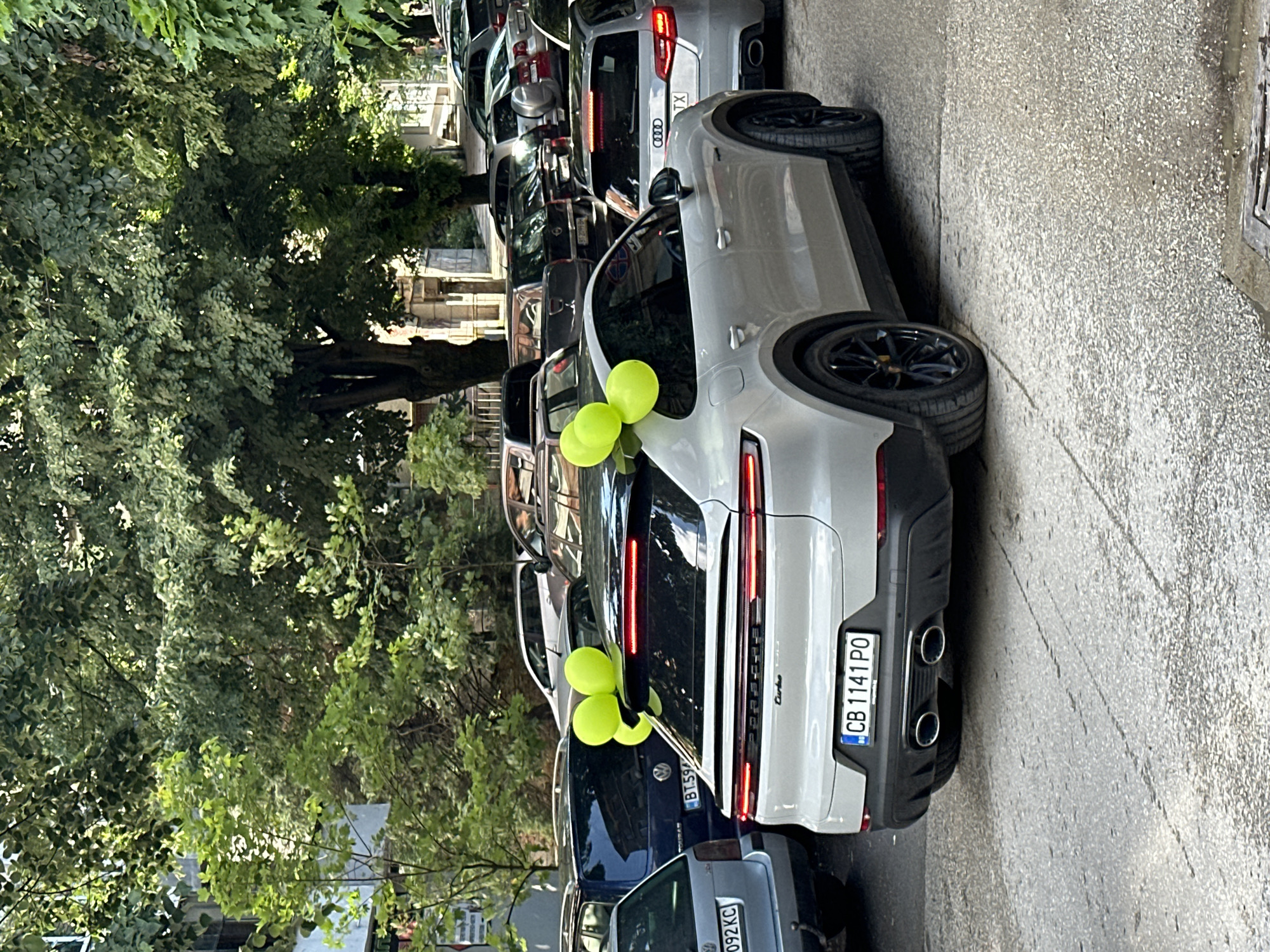 The image depicts a street in Veliko Tarnovo, Bulgaria, densely packed with parked cars. The focus is on a light-grey Porsche with yellow balloons tied to its rear, indicating a celebration or special event. The car's license plate reads "CB 1141 PO." Other cars are parked tightly around it, showing a mix of models and colors. The environment includes trees lining the street, suggesting an urban, residential area. The image orientation is sideways. There are no visible people.