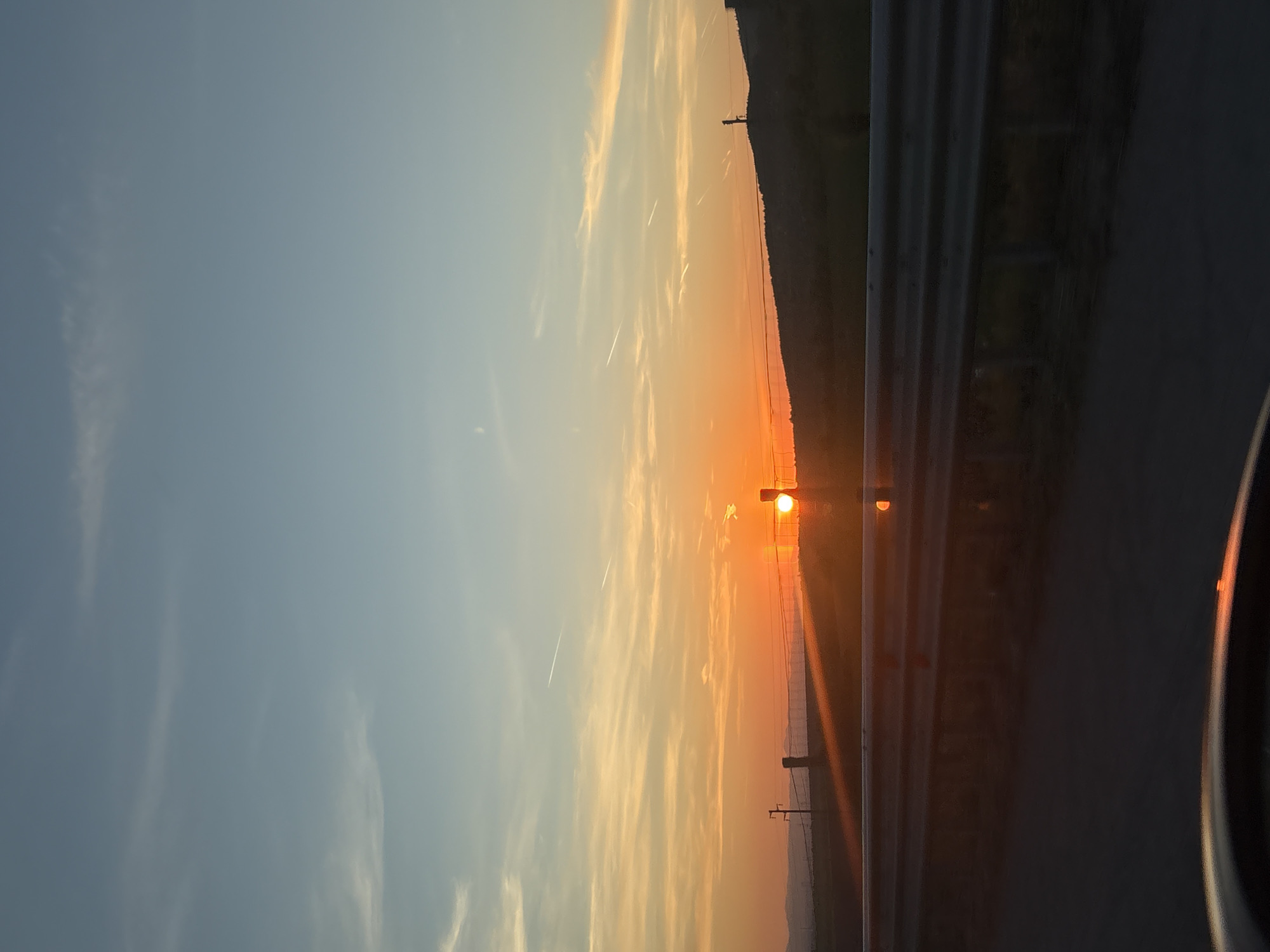 Captured from inside a vehicle, a sunset dominates the sky near Eleshnitsa, Bulgaria. To the right, a roadside guardrail is visible, partly obstructing the view of a hill in the distance. Power lines and utility poles punctuate the landscape below the setting sun. Streaks of vapor trails cut across the sky, contrasting with the gradient of blue to orange hues. Parts of the vehicle's interior are visible on the right.