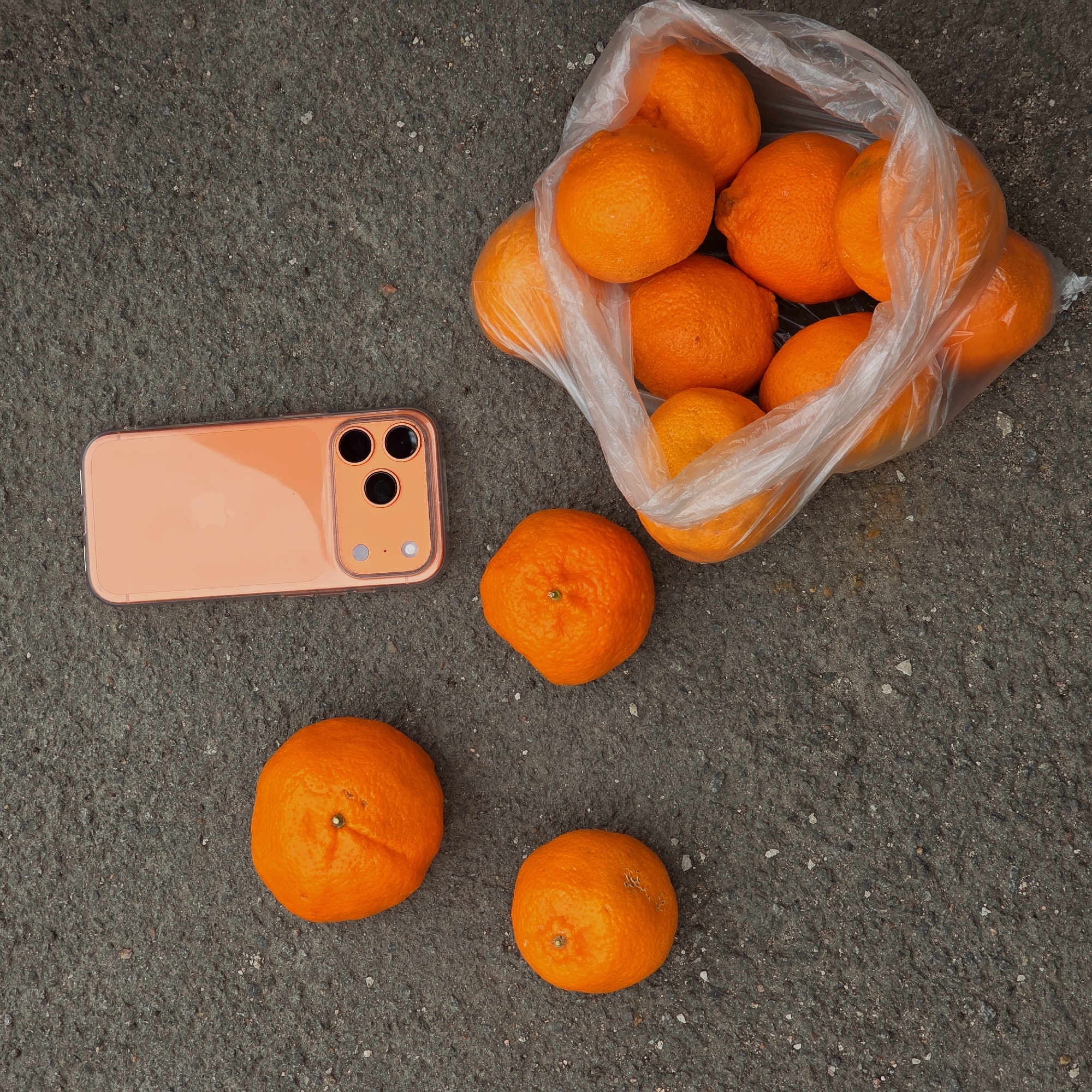 The image shows a bag of oranges and a smartphone on a gray asphalt surface. There are approximately 10 oranges in a clear plastic bag. Next to the bag is a smartphone with an orange case. Three loose oranges are scattered near the bag and smartphone. The scene appears to be outdoors, possibly on a street or sidewalk. There are no people in the frame. There is no information to suggest the location is Saint Petersburg, Russia.