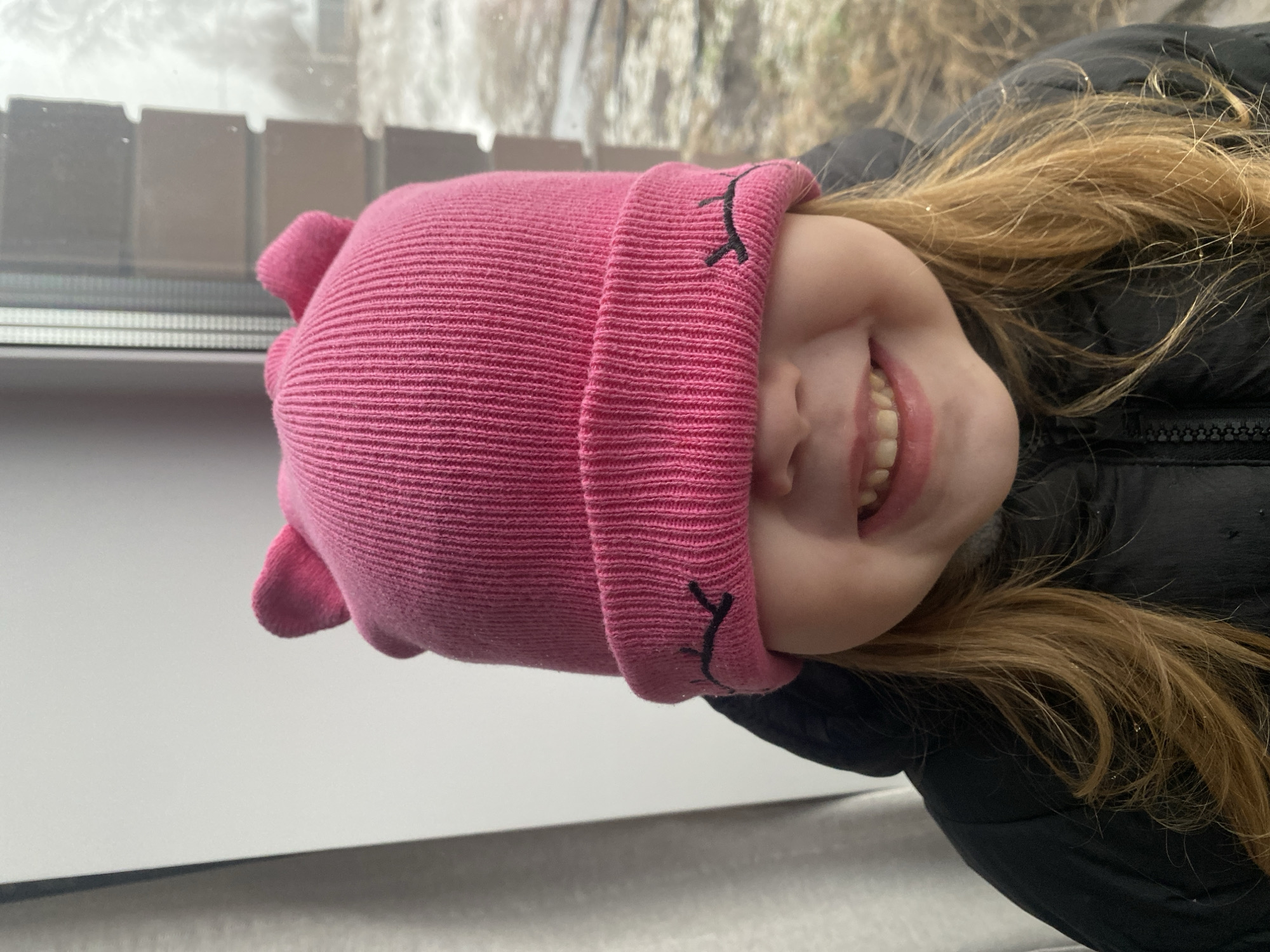 A young girl wearing a pink knit hat with embroidered eyelashes is smiling and looking towards the viewer. She is wearing a black puffer jacket. In the background, a window with several panes is visible, showing an outdoor scene with snow and blurred vegetation. The image was taken indoors. Without additional information or context, I cannot confirm the location is Izhevsk, Russia.