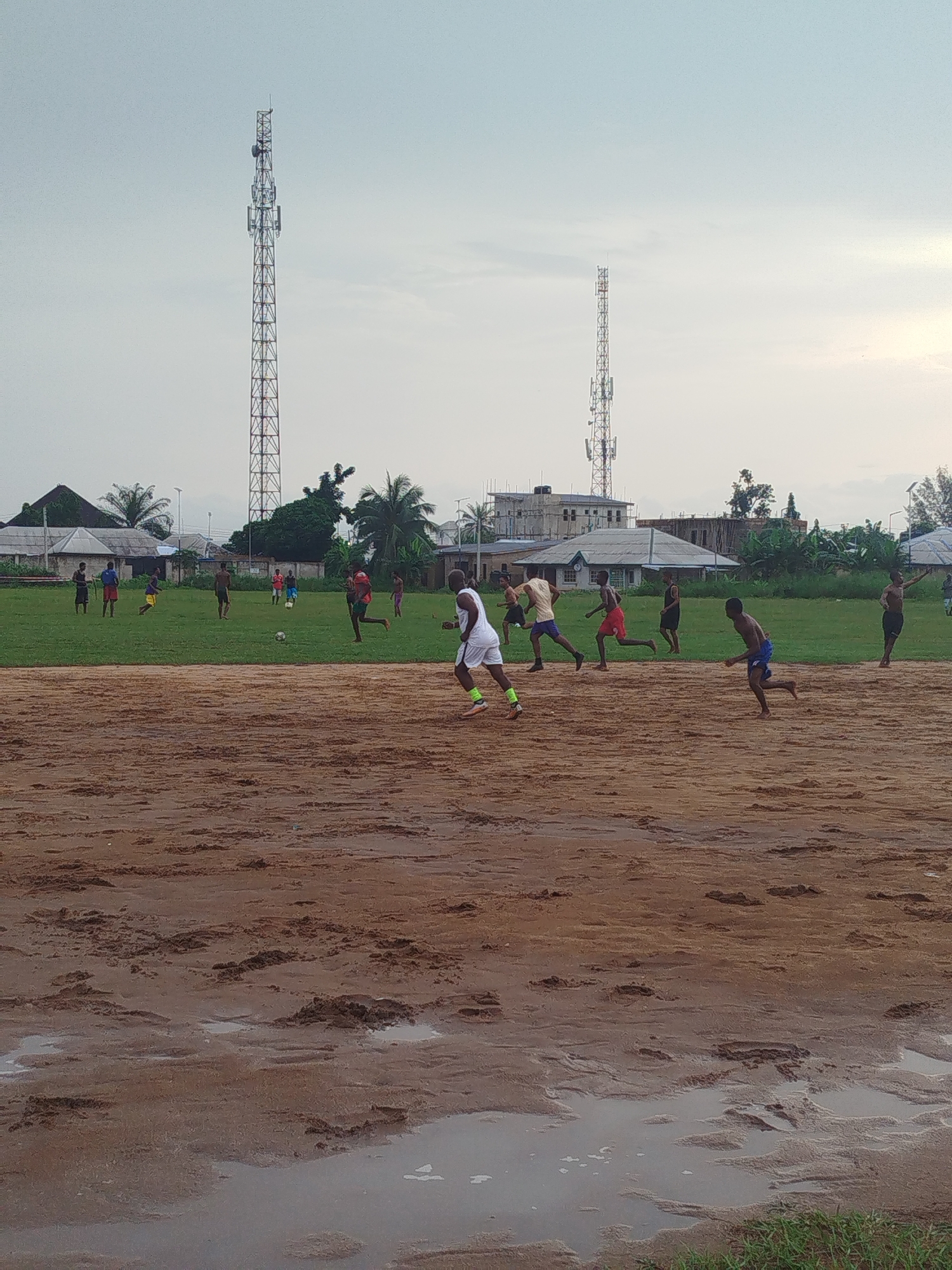 In Ikot Mbonde, Nigeria, a group of young men are playing soccer on a field. The foreground is muddy with standing water. The playing field is partly green, partly brown dirt. Two communication towers are visible in the background along with houses and trees. The sky is overcast. The players are in various stages of running.