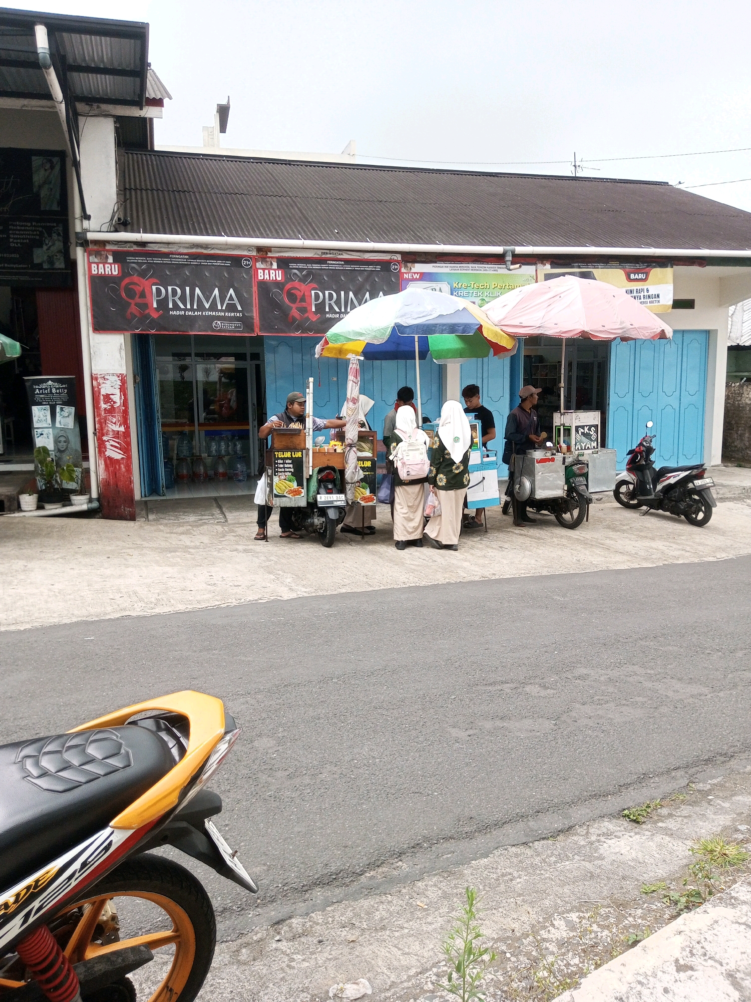 In the city of Krasak, Indonesia, a street scene unfolds with a foreground occupied by the rear section of a parked motorcycle. The motorcycle features a black seat with a textured pattern, a bright yellow handle, and orange-spoked wheels, with visible red suspension springs. The background depicts a row of shops with dark gray corrugated roofs. A prominent banner displaying "PRIM A" in red and black lettering with the tagline "HADIR DALAM KEMASAN KERTAS" is visible. To the right, another banner announces "KRE-TECH PERTAMA" and "DARI KRETEK KRETEK KRETEK." Underneath these banners, blue shop doors are visible. In front of the shops, several individuals are gathered around food carts. One cart displays signage for "TELUR LUR." Three young women, two wearing hijabs and one with a backpack, are standing near a food cart. Another man, wearing a cap and a dark shirt, is attending to a food cart. A third man, wearing a blue shirt and a cap, stands next to a food cart. Two motorbikes are parked near the food carts, one white and red, and another dark colored with a silver box attached. Umbrellas, one pink and white and another blue and green, are set up over the food carts. The environment appears to be a commercial street with a paved road and sidewalk.