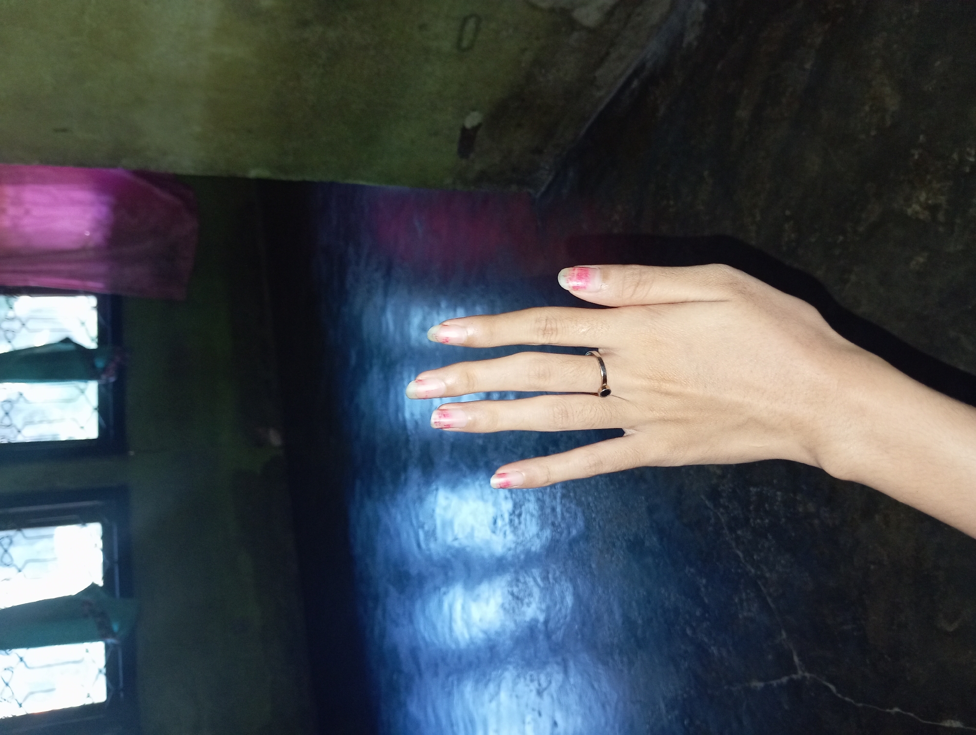 A female hand with painted fingernails is extended forward, displaying a ring on the ring finger. The background is dark and appears to be an interior space with windows and a textured wall. The floor is reflective and shows some light patterns. The overall lighting is dim, with some light filtering through the windows. The location is described as being in the city of Rejosari, Indonesia.