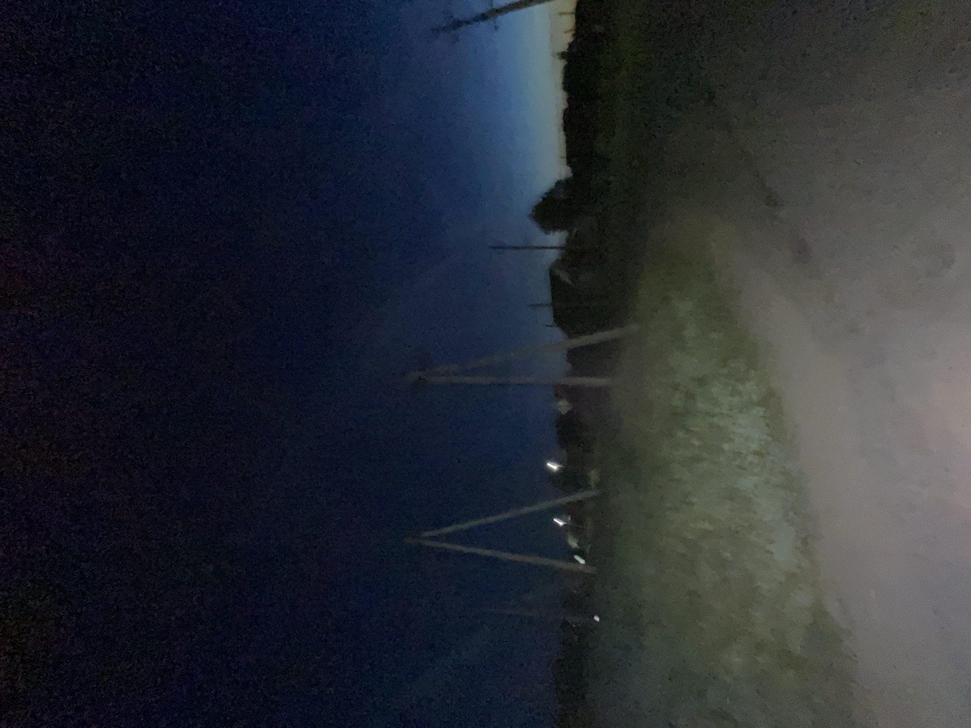 The image shows a dirt road in a rural area at night. There are power lines visible on the left and right sides of the road, as well as in the background. Some houses and trees are silhouetted against the dark blue sky. The overall impression is one of a quiet, rural landscape at dusk or dawn. The location is identified as Krasnye Baki, Russia.
