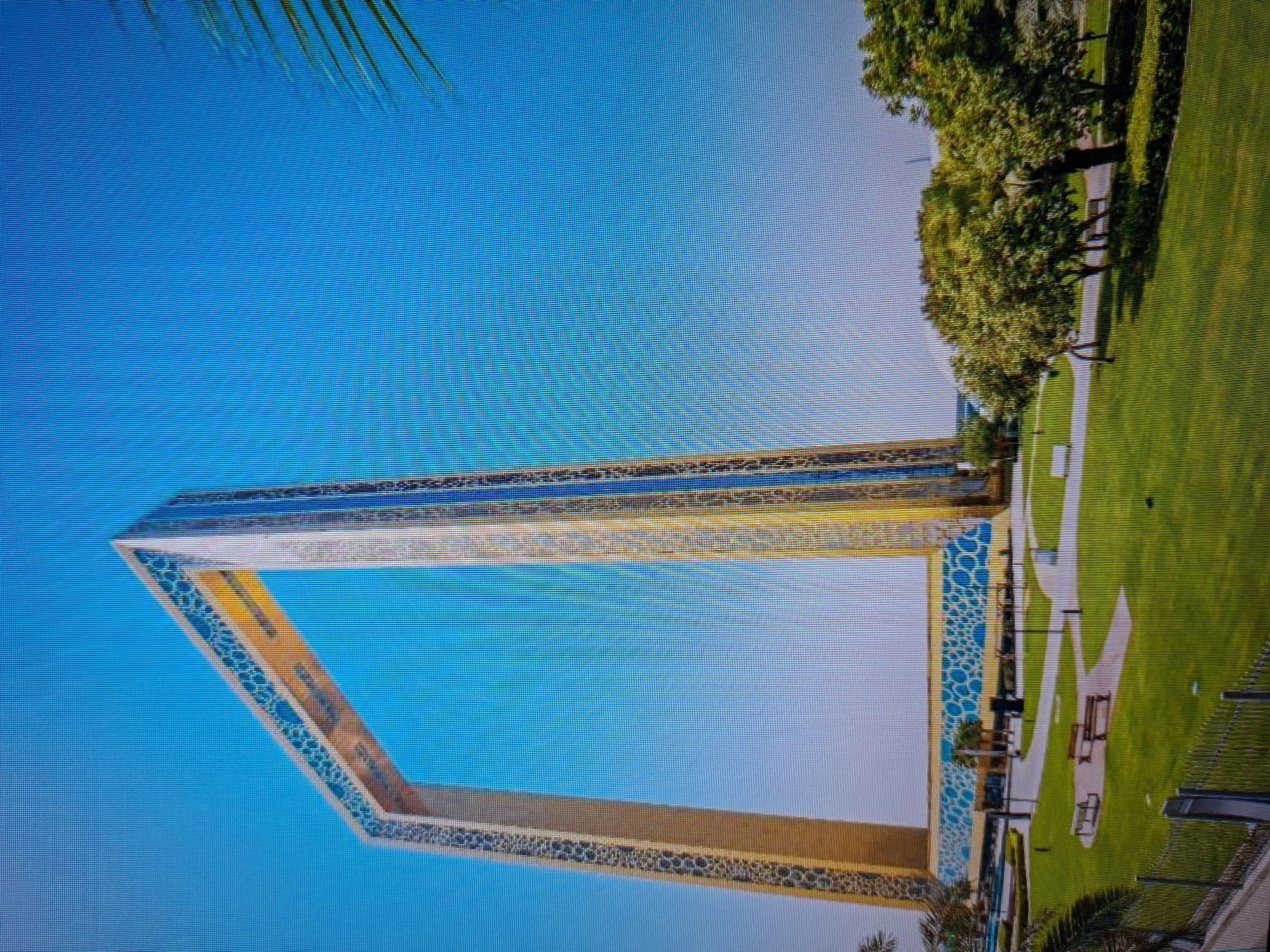Here is a forensic description of the image:
The image shows the Dubai Frame, a large rectangular structure in Dubai, United Arab Emirates. It is positioned against a clear blue sky with a few palm fronds visible in the upper left corner. To the right of the frame, there is a park with green grass, trees, and walkways. Benches are visible along the walkways. The frame itself is decorated with a geometric design along its edges, and its interior creates a window onto the city skyline. The image appears to be taken from a low angle, emphasizing the height and scale of the Dubai Frame.