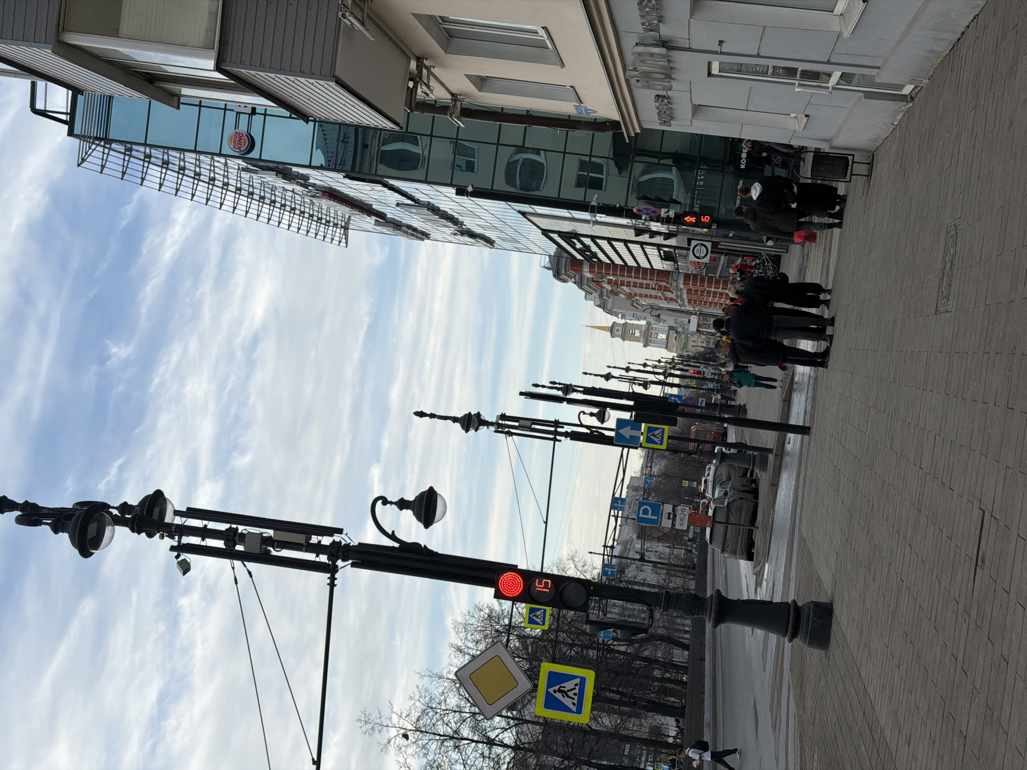 The image depicts a street scene in Perm, Russia. Pedestrians are gathered on the sidewalk near a building with mirrored glass reflecting the sky and clouds. A traffic light displays a countdown indicating "5" seconds, positioned on a lamppost with pedestrian crossing signs. Further down the street are multiple lampposts and street signs. Buildings are visible in the background, including one with red brick accents and a clock tower in the distance. Cars are parked on the side of the street. The sky is partially cloudy.