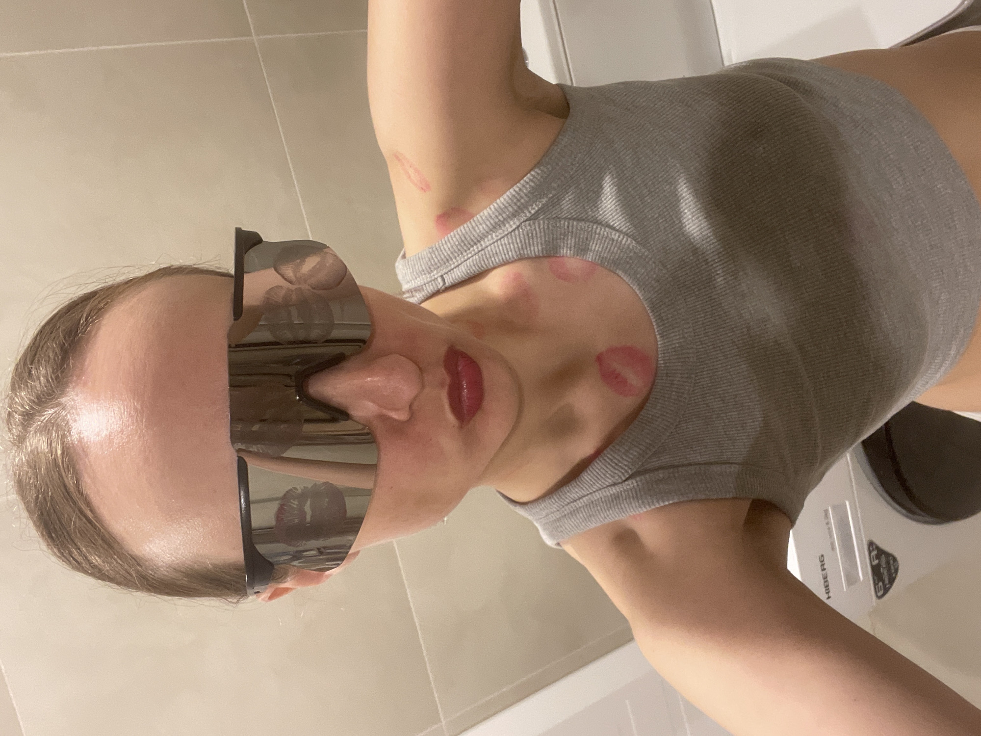 A woman with fair skin and light brown hair is depicted wearing large, dark sunglasses and a gray tank top. She has several lipstick marks on her neck and chest. The background appears to be a tiled bathroom with a washing machine visible. The location is unspecified.
