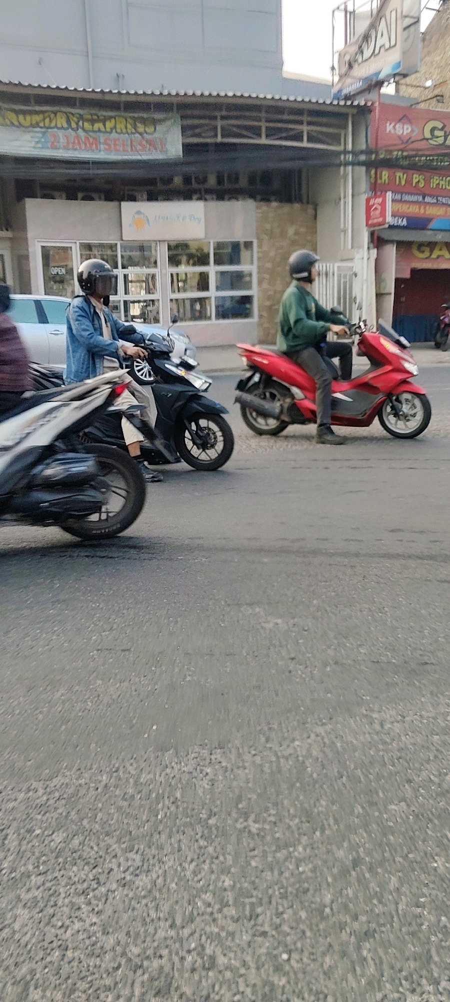 In Mekarmukti, Indonesia, a street scene unfolds with several motorbikes and individuals. A man wearing a blue denim jacket and a black helmet with a dark visor rides a black motorbike. Beside him, on the right, another man in a green long-sleeved shirt and a black helmet is on a red motorbike. In the foreground, a portion of a white and black motorbike is visible. The background shows buildings with shop signs, including one that says "LAUNDRY EXPRESS 2 JAM SELSA." The environment appears to be a commercial area with asphalt road surfaces. The lighting suggests it is daytime, with the sun casting shadows.