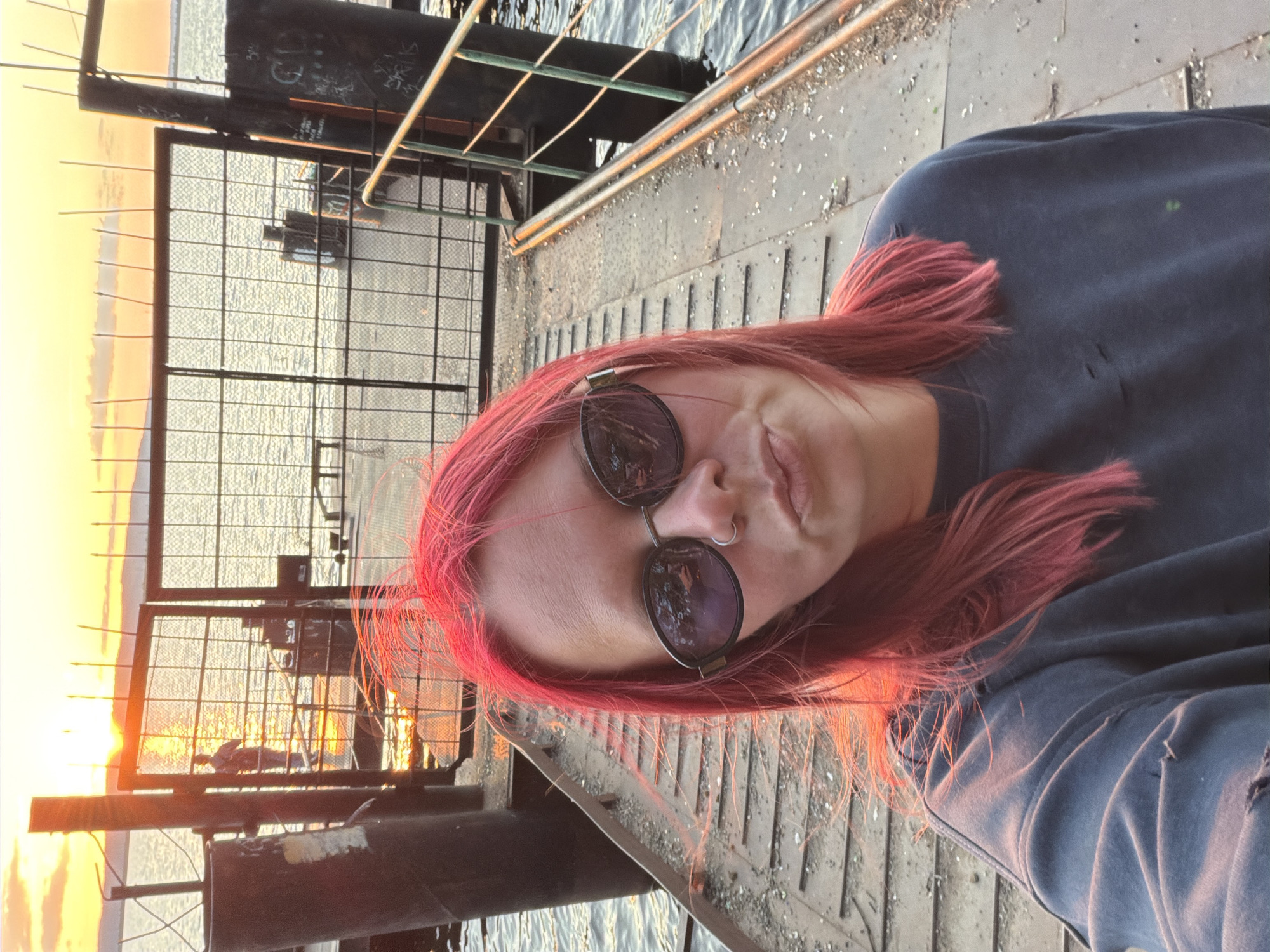 The image shows a person with bright pink hair wearing sunglasses and a dark grey sweatshirt with ripped details, taking a selfie on a dock at sunset. The person is leaning into the camera with their lips pursed. Behind them, the sun is setting over a body of water, casting a warm orange glow. The dock is made of metal and wood, with railings on the right side. There are bollards and metal fencing visible. In the distance, a person can be seen standing on a structure over the water, silhouetted against the sunset. The overall scene suggests a casual, outdoor setting by the water during the golden hour.