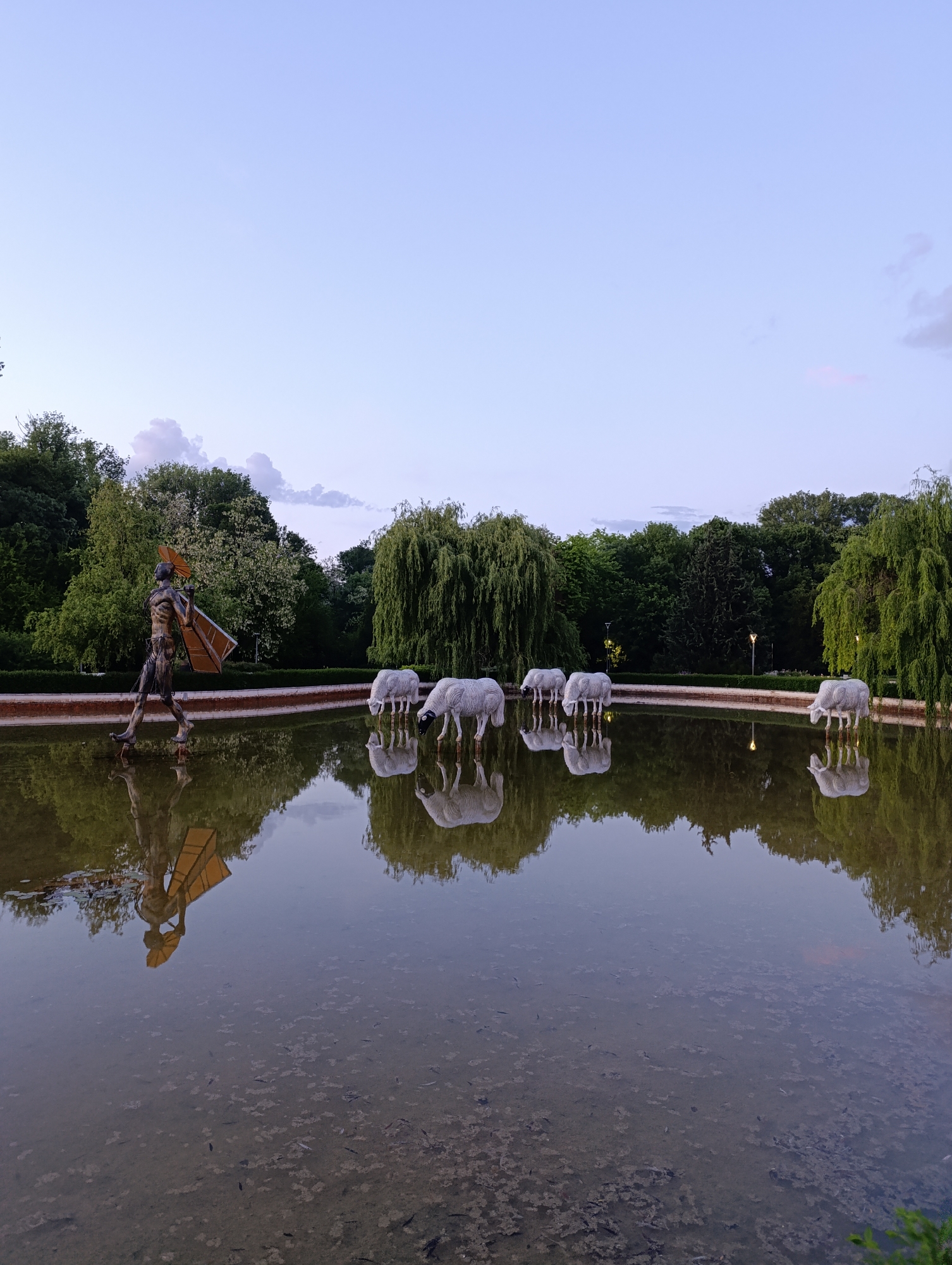 The image shows a pond in a park setting, likely taken at dusk. In the foreground, a bronze statue of a figure carrying a rectangular object is partially submerged in the water. The statue's reflection is visible in the pond. Further into the pond, there are five white sheep sculptures with their heads lowered, as if drinking water. Their reflections are also visible in the pond. The pond is surrounded by greenery, including trees and bushes. In the background, there is a row of trees and a clear sky with a faint hint of sunset.
This scene is likely located in Borisova Gradina (Boris' Garden), in Sofia, Bulgaria, specifically near the Ariana Lake (Ariana езеро). The statues of the sheep are a known feature of this park area.