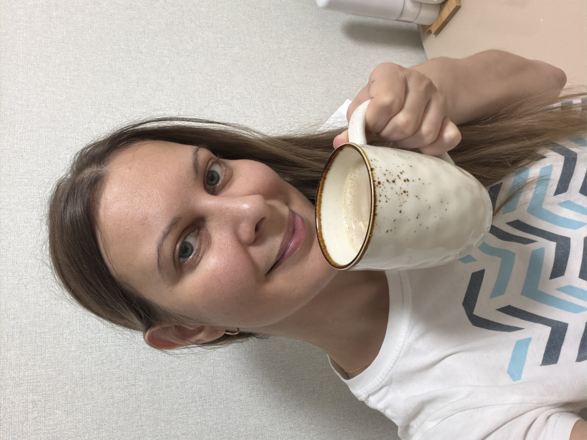 A fair-skinned young woman with shoulder-length brown hair is holding a partially filled mug, likely containing a latte or similar coffee drink. She is wearing a white t-shirt with a geometric pattern in light blue and dark gray. The background consists of a plain, light-colored wall. The photo does not provide any context to ascertain if this photograph was taken in Izhevsk, Russia.