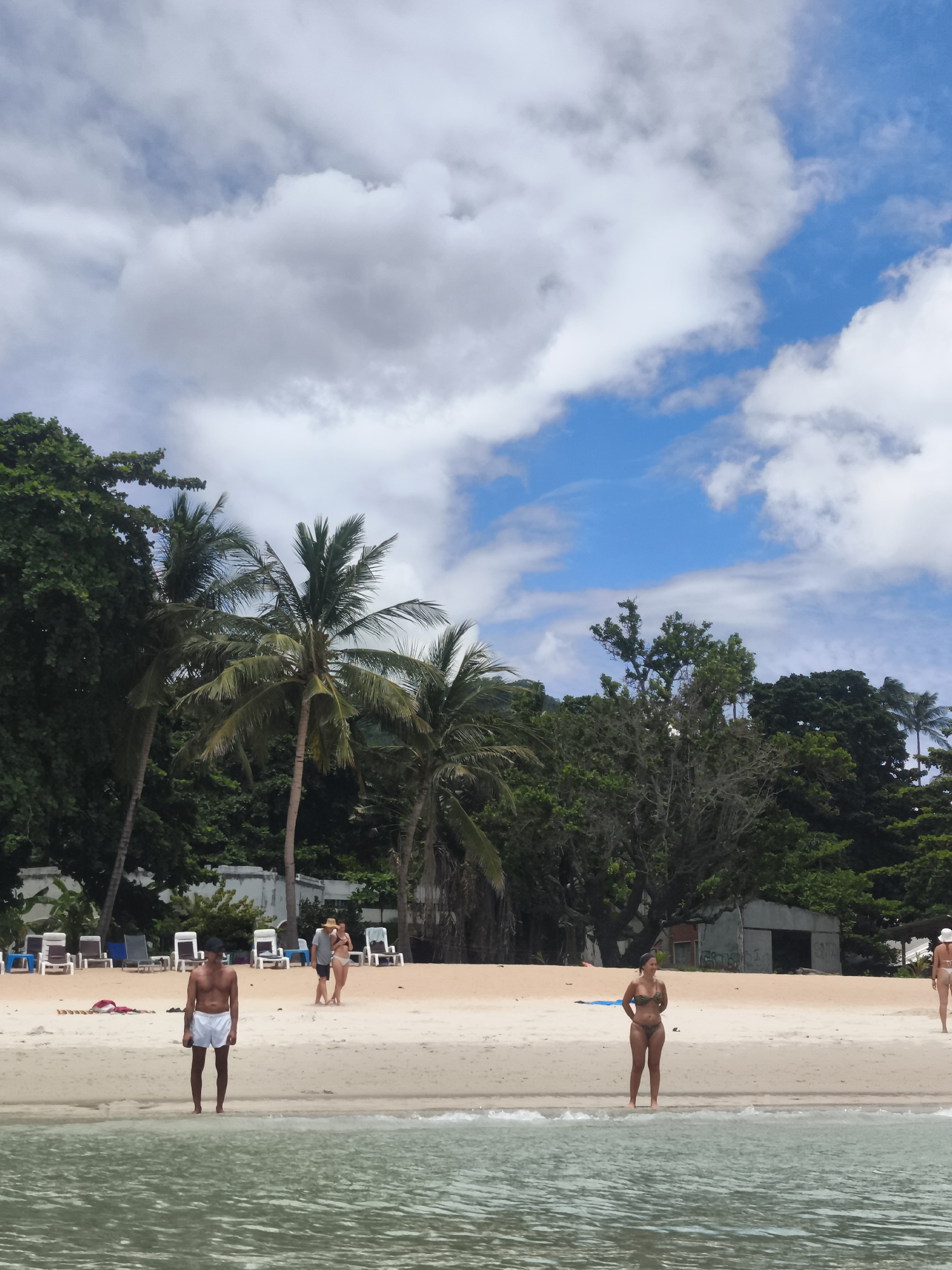 This image depicts a scenic beach in the Ko Samui District of Thailand. The foreground shows calm, clear ocean water. A man in white shorts stands near the water's edge, facing the ocean. Further back on the sandy beach, a woman in a patterned bikini stands facing left. In the background, several lounge chairs are set up on the sand, with a line of lush green trees and palm trees forming a dense backdrop. A few people are visible walking and standing on the beach. The sky above is a mix of blue and white clouds.