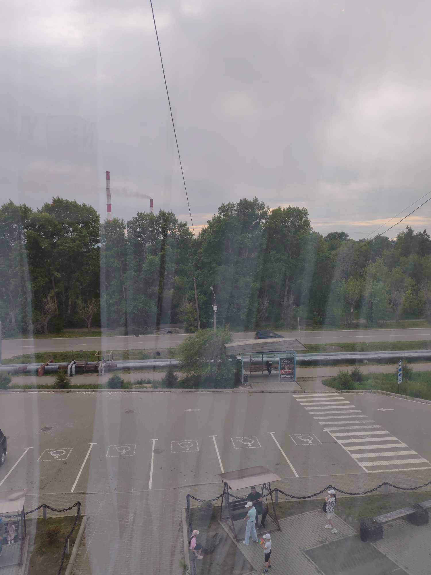 The image shows a view from above, looking out over a road and a parking lot. In the distance, two tall industrial smokestacks with red and white bands emit light smoke into an overcast sky. A line of dense green trees borders the road.
In the foreground, a paved parking lot has several marked parking spaces, four of which are designated for disabled individuals, indicated by the International Symbol of Access. A pedestrian crosswalk is clearly marked, leading across the parking lot towards a bus stop shelter. Several people are visible in the scene: on the swings under a canopy, standing near the bus stop, and walking in the parking lot. A bench is situated in the lower right corner of the image. A parked car is partially visible on the left side of the parking lot.
The scene appears to be located near an industrial area, suggested by the smokestacks, and a public space with a road, parking, and public transport facilities.