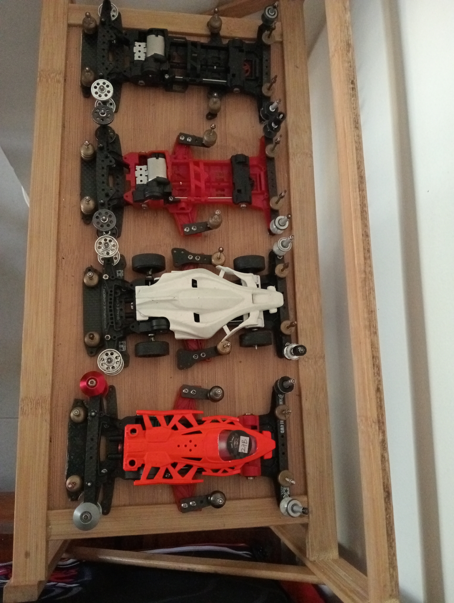 The image shows four slot cars lined up vertically on a wooden surface. The top car is black with silver wheels. The second car from the top is red with black wheels and has a white top. The third car is white with black wheels and silver accents. The bottom car is orange with black wheels and red accents. The cars are positioned on a light-colored wooden shelf, possibly part of a display unit. The background is a white wall on the right and a blurred dark area on the bottom. No people are visible in the image. The scene appears to be indoors.