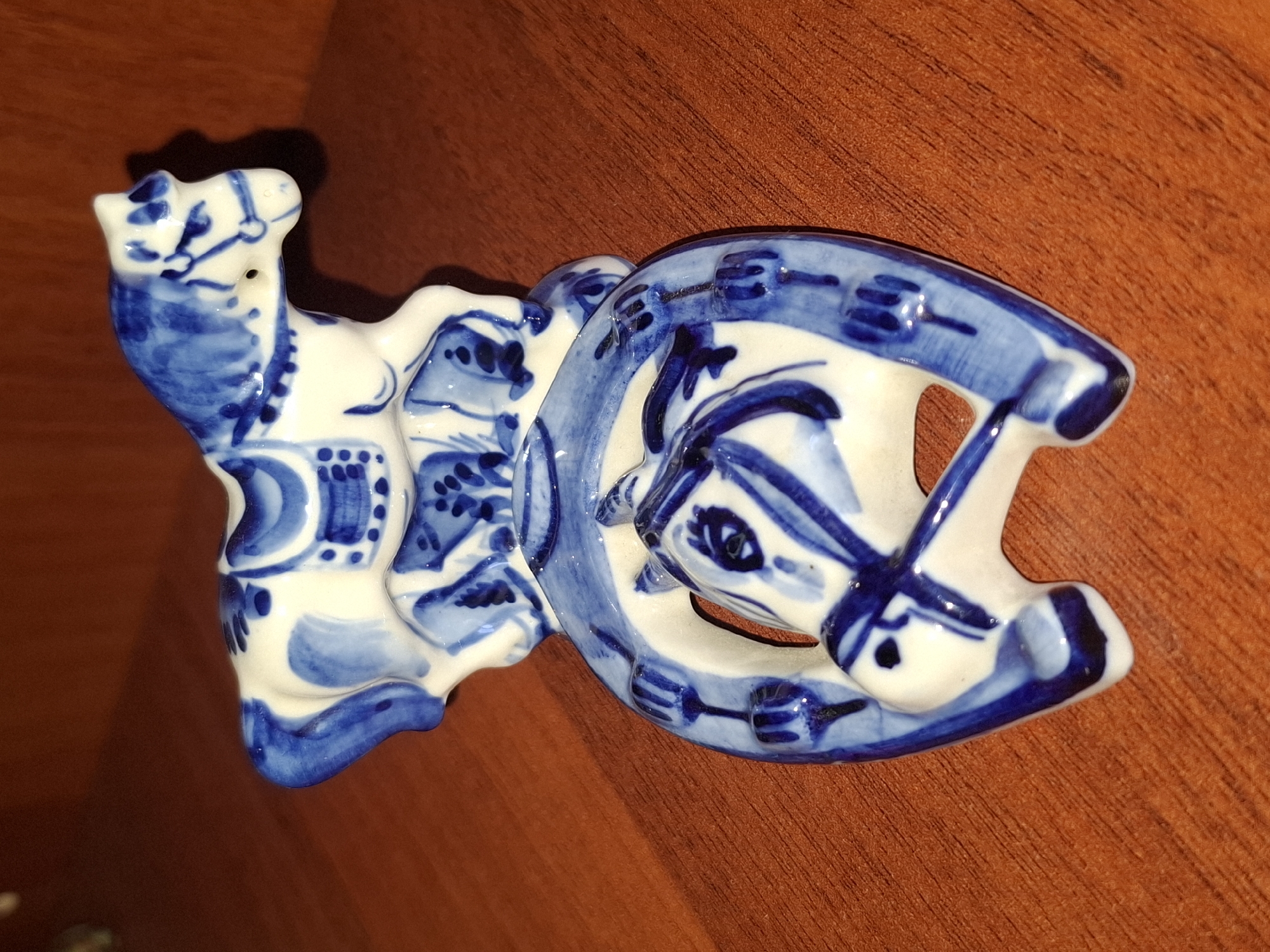 A white porcelain figurine with blue painted designs is placed on a brown wooden surface. The figurine depicts a horse standing on top of a horseshoe, with another horse's head emerging from the horseshoe. The horse on top has a saddle and bridle painted on it. The horseshoe is decorated with blue patterns and has a horse's head as part of its design. The background is a wooden surface, possibly a table or shelf. The lighting casts a shadow from the figurine onto the surface. There is no indication of the city of Almaty, Kazakhstan, or any other specific location.