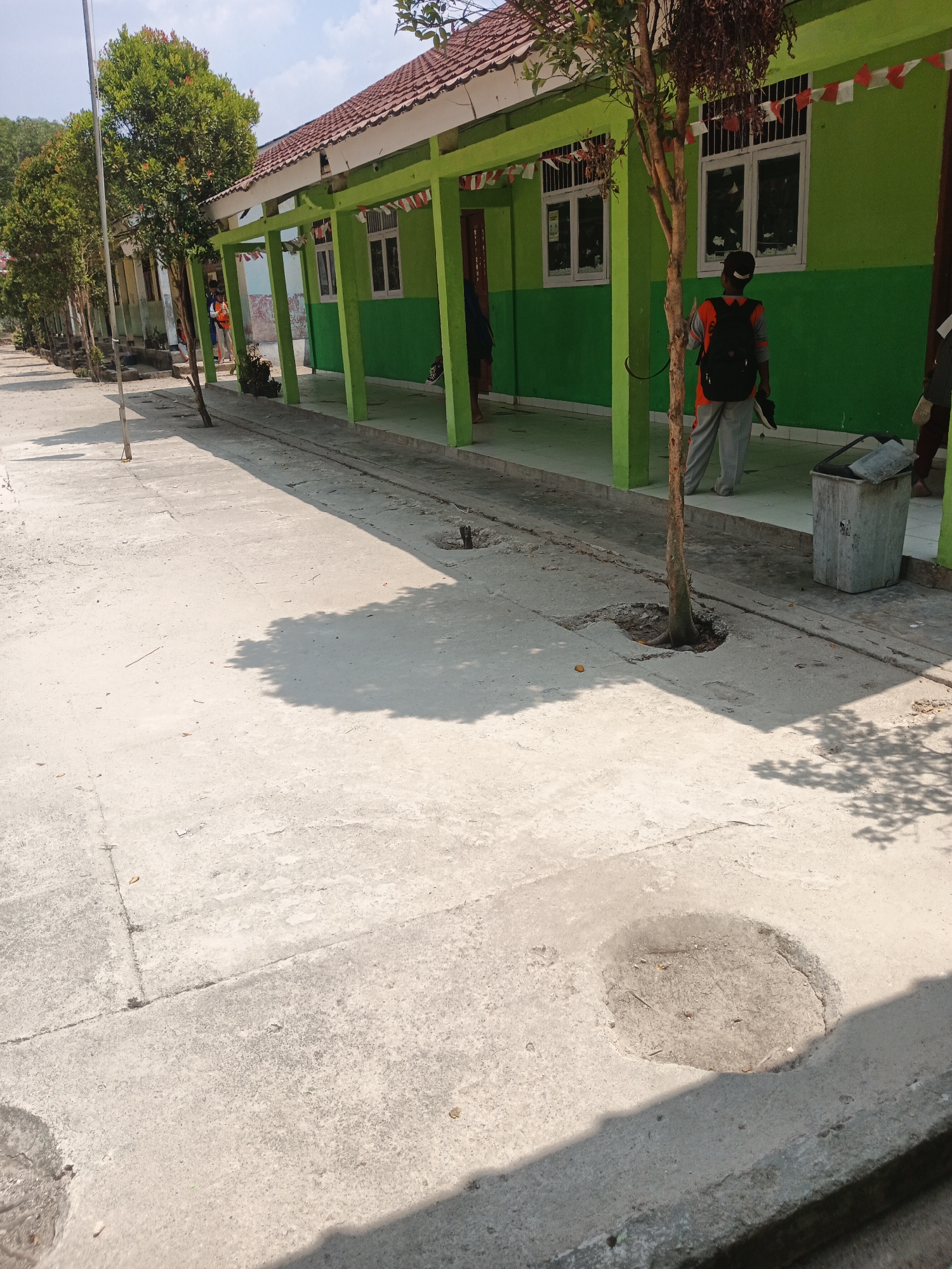 In Rejo Basuki, Indonesia, a sun-drenched outdoor pathway runs alongside a green school building. The pathway, made of concrete, shows signs of wear with several small holes and cracks. A young boy, seen from behind, is standing on the pathway, wearing a backpack and grey pants. He is holding a pair of shoes. Next to him, a large, weathered metal trash can stands. The school building features a red-tiled roof, white trim, and green walls. Red and white Indonesian flags are visible decorating the building's facade. Small trees line the path, casting shadows on the concrete. The sky above is clear and blue.