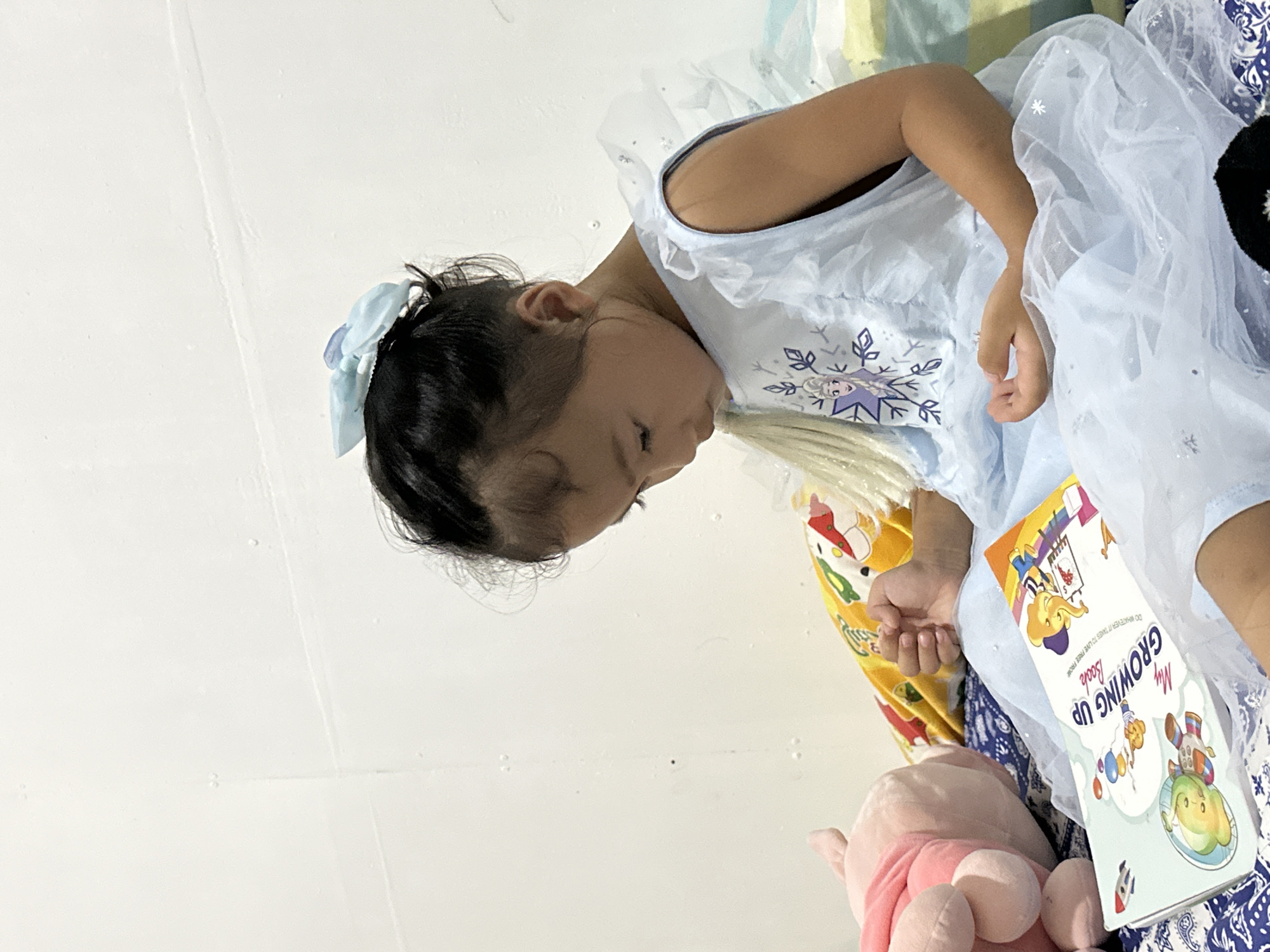 A young Filipina girl, adorned in a light blue, sleeveless dress with a Frozen Elsa graphic and sparkly accents, sits on a bed. She has dark hair tied with a light blue bow. The girl is looking down at an open book titled "My Growing Up Book," which features colorful cartoon illustrations. To her left, a pink stuffed animal is visible. The background is a plain white wall. The scene appears to be indoors, likely in a residential setting in Davao City, Philippines.