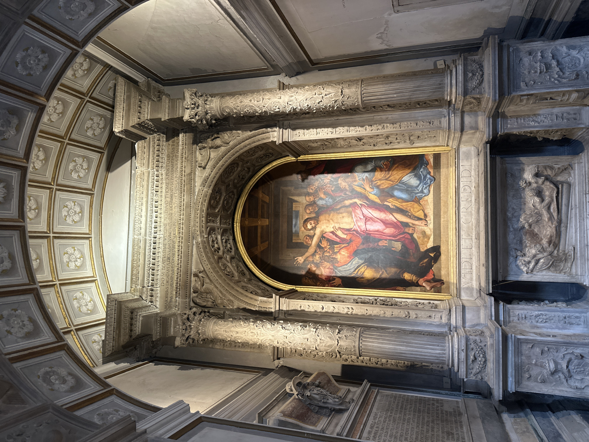 The image shows an altarpiece depicting the Crucifixion of Jesus Christ, located in the Cappella Sansevero in Naples, Italy. The painting is centrally positioned within an ornate architectural frame featuring Corinthian columns and intricate sculptural details. Above the painting, an inscription is visible, likely part of the chapel's dedication or a memorial. The ceiling is adorned with decorative coffers and floral motifs. The environment appears to be a dimly lit, historical interior space, possibly a church or chapel, with stone walls and decorative elements characteristic of Baroque architecture. A sarcophagus-like structure is visible at the base of the altarpiece, featuring relief sculptures. The overall impression is one of solemnity and artistic grandeur.