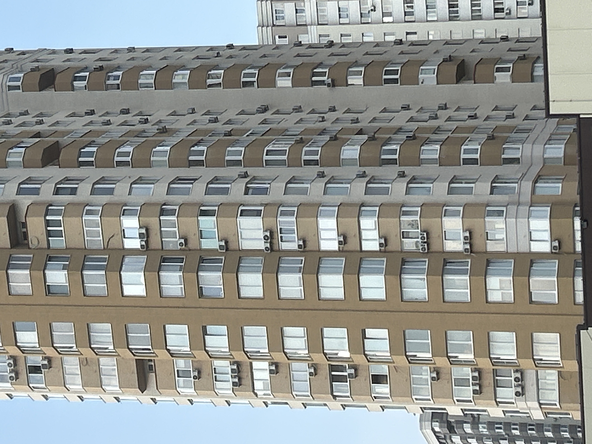 The image captures a close-up, low-angle view of a tall, multi-story residential building in Brovary, Ukraine. The building features a predominantly beige facade with numerous windows and balconies. The architectural style suggests a modern, mid-to-late 20th-century construction. Air conditioning units are visible mounted on the exterior walls below many of the windows. The building's facade is segmented by vertical lines, creating a series of vertical bays, and horizontal bands of balconies with curved railings. The sky is a clear, pale blue, indicating fair weather. To the right, another, lighter-colored residential building of similar height is partially visible, with a repeating pattern of windows. The perspective emphasizes the verticality of the structures, with the top of the building extending out of frame. There are no people or vehicles visible in the image.