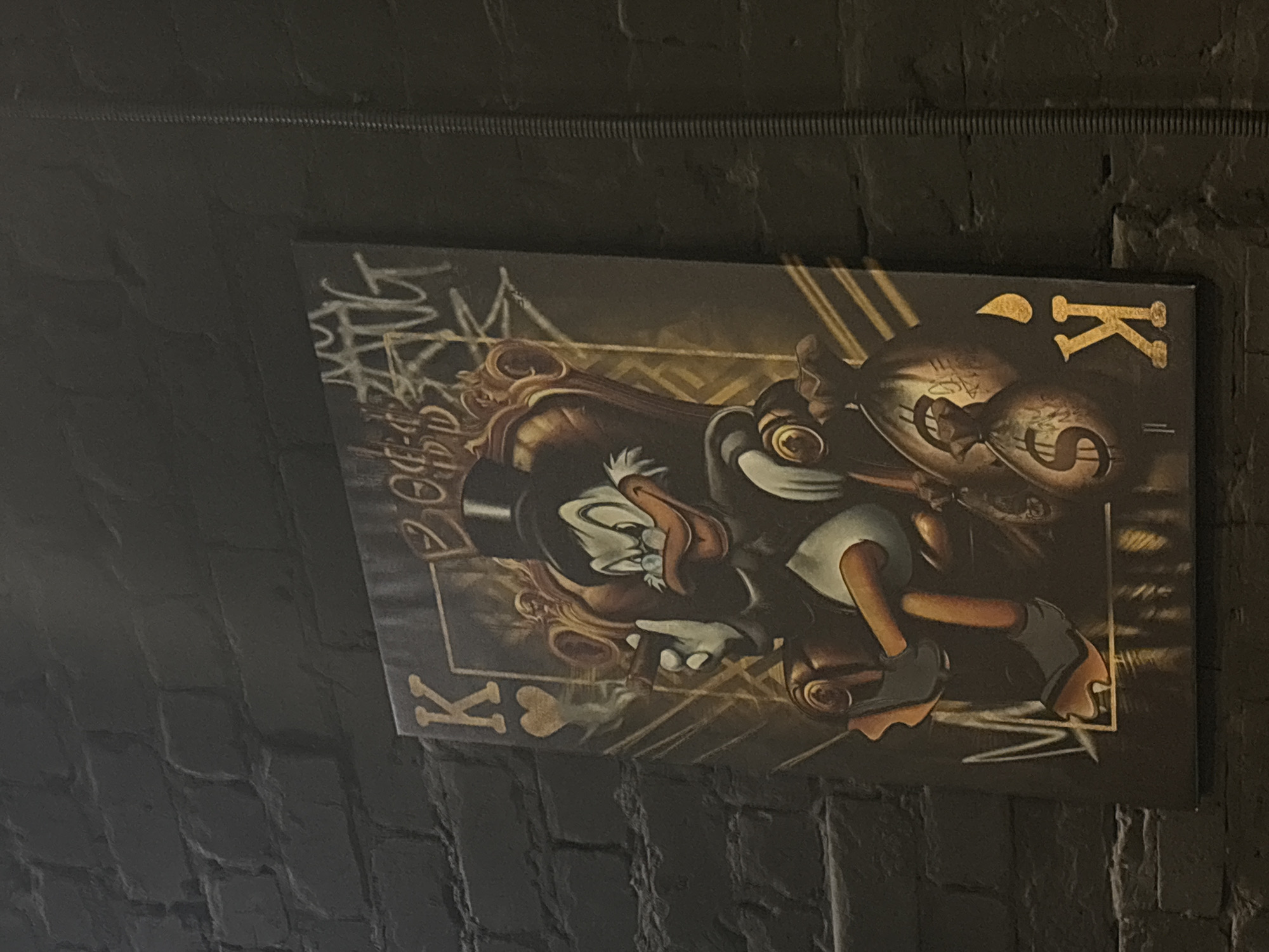 The image displays a painting of Scrooge McDuck dressed in formal attire and a top hat, seated on a golden throne. He is holding a cigar in his left hand and appears to be looking to his right with a confident expression. Two money bags with dollar signs are placed at his feet, and the painting features a playing card motif, with a "K" symbol visible in the upper left and lower right corners. The painting is mounted on a dark brick wall, which shows signs of weathering and texture. The lighting in the scene is dim, highlighting the painting and the dark background. The photograph appears to have been taken indoors, possibly in a bar or a similar establishment. This image is located on a brick wall in Chaikovsky, Russia.