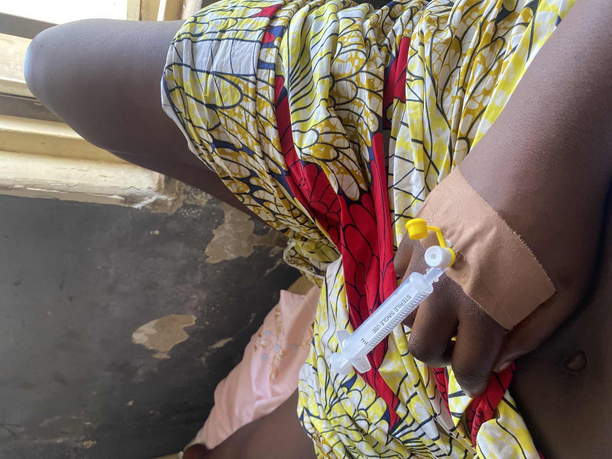 A person is seen sitting, their hand secured with a tan bandage and connected to a clear plastic syringe with a yellow and white cap. The syringe is labeled "STERILE SINGLE USE." They are wearing a colorful garment featuring yellow, white, red, and blue patterns. A pink garment is partially visible in the background. The setting appears to be indoors, possibly a building in Rido, Nigeria, based on the prompt's instructions, with what appears to be a concrete floor visible.
